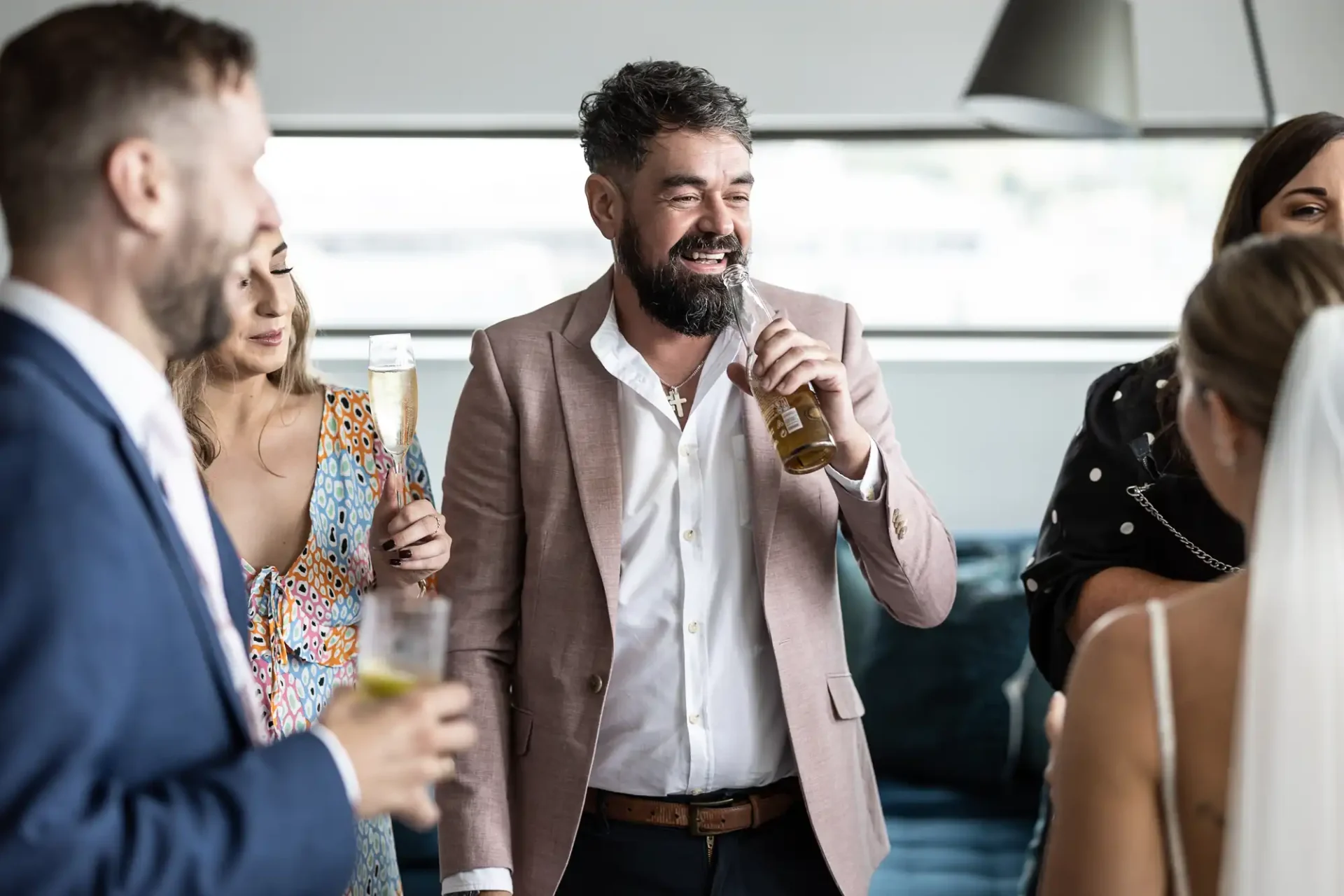 Man in a beige blazer laughing and holding a beer, enjoying a conversation with friends at a social gathering.