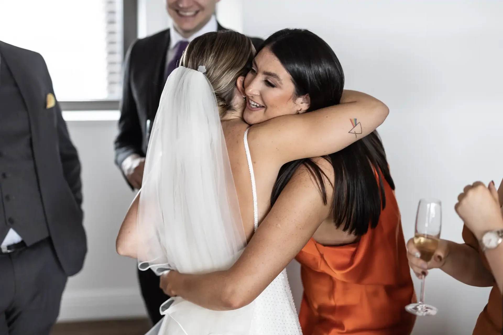 Bride in white dress embracing a woman in an orange dress at a wedding, with guests holding champagne around them.