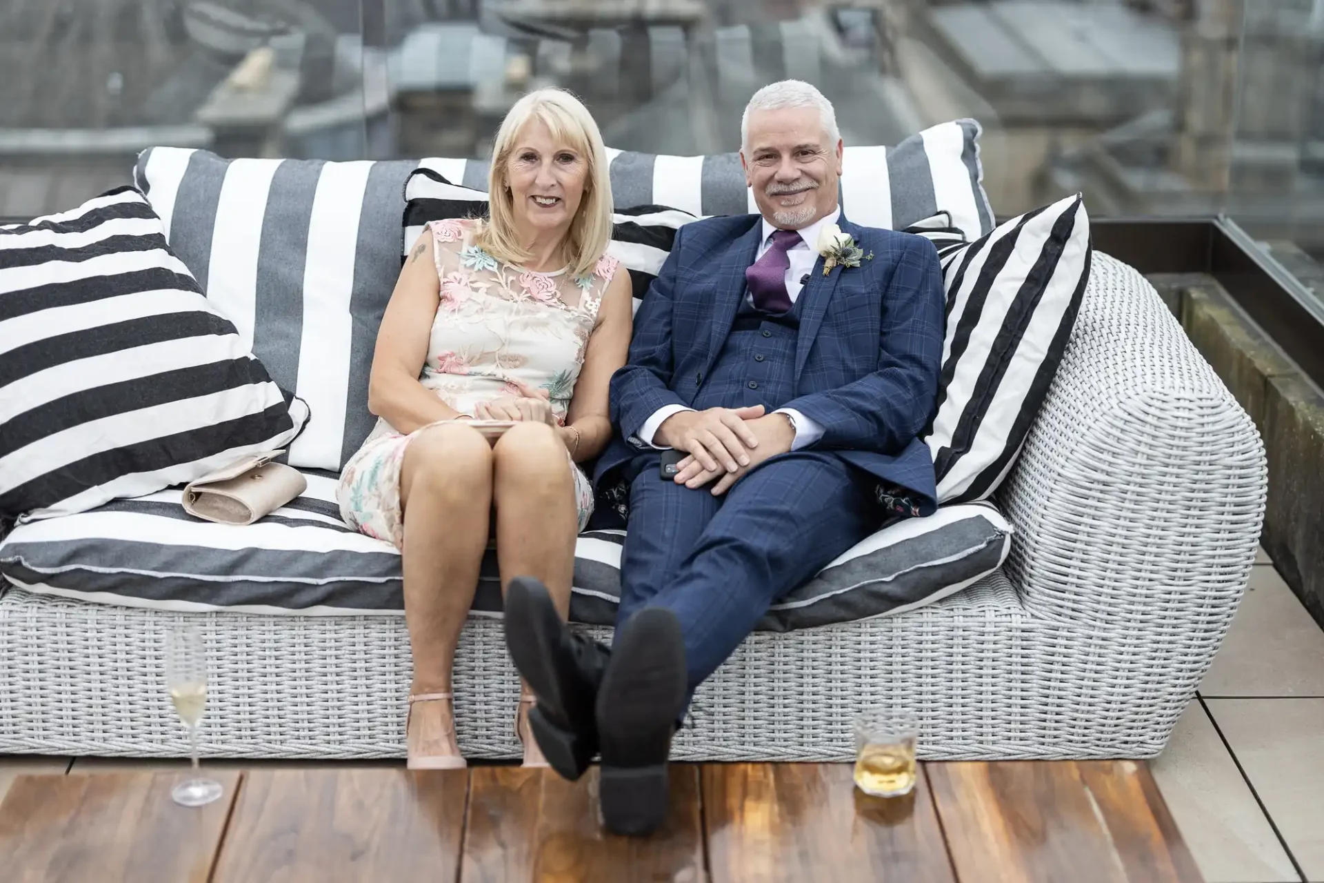 An older couple dressed in formal attire smiles while sitting closely together on a striped sofa, with drinks beside them on an outdoor patio.