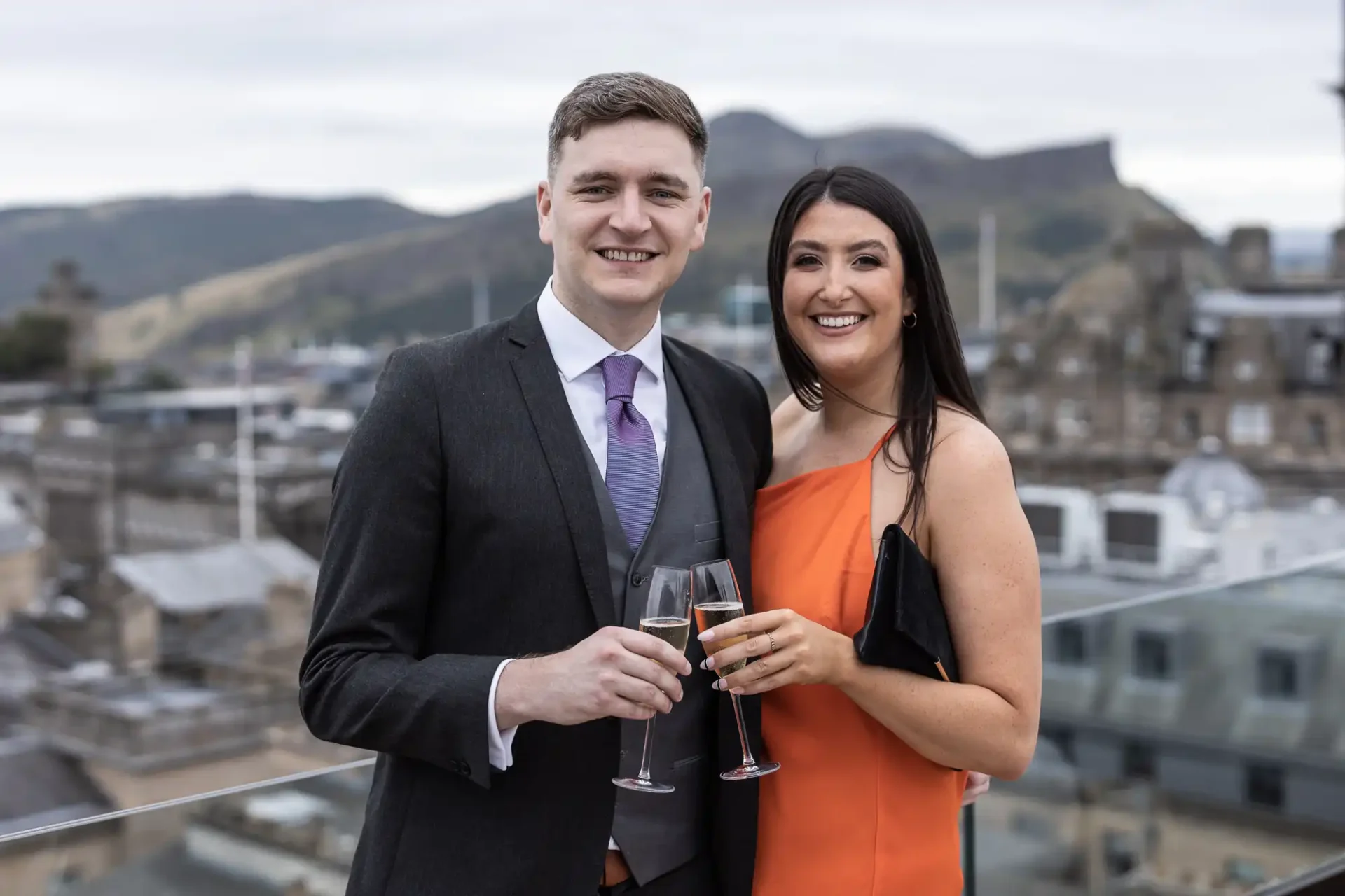 A man and a woman smiling, holding champagne glasses on a rooftop with a cityscape and hills in the background.