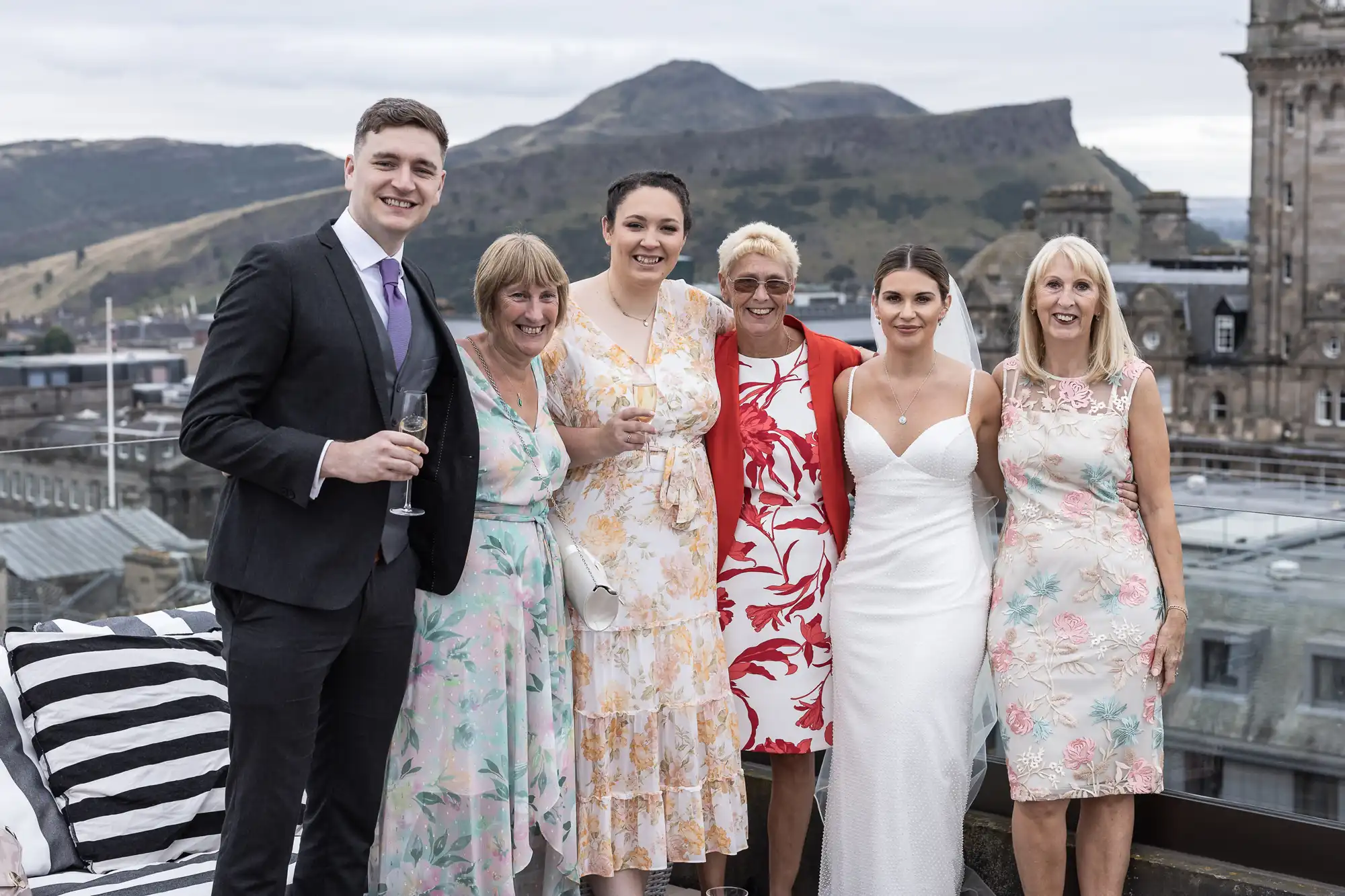 Group of six people dressed in formal wear, holding glasses and smiling, with a cityscape and a hill in the background.