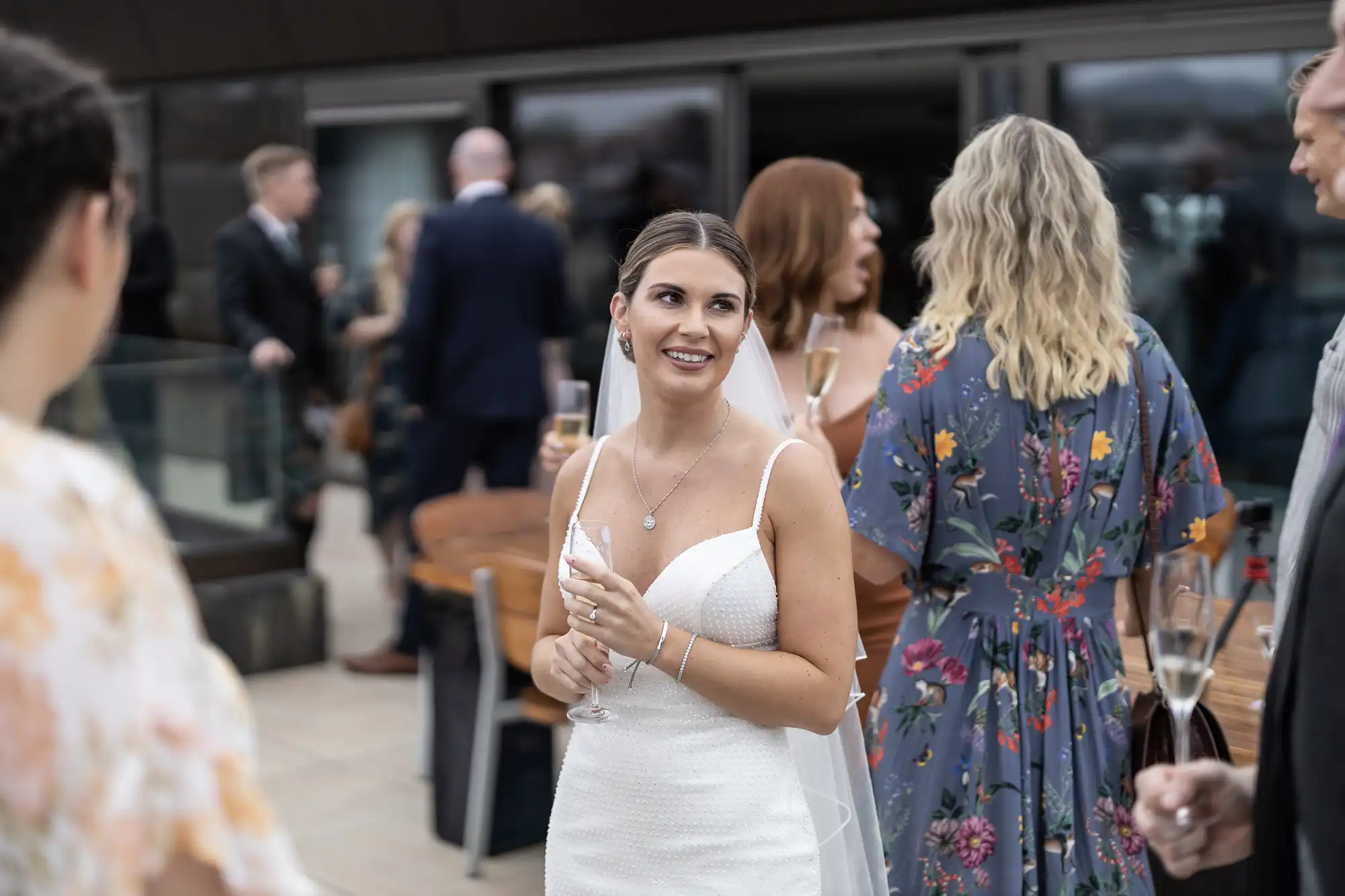 A woman in a white dress holding a champagne glass at a social event, smiling and engaging in conversation with other guests.