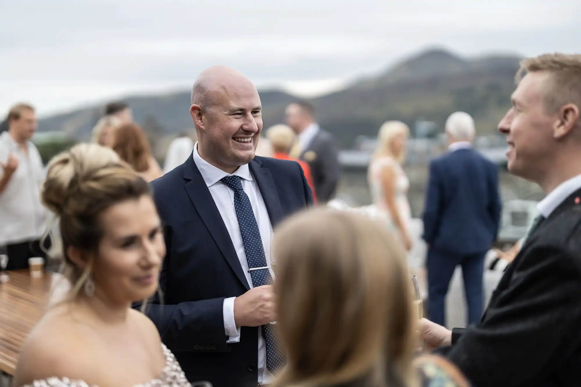 A man in a suit laughs while talking with another man at an outdoor social event, with people in formal attire around them and hills in the background.