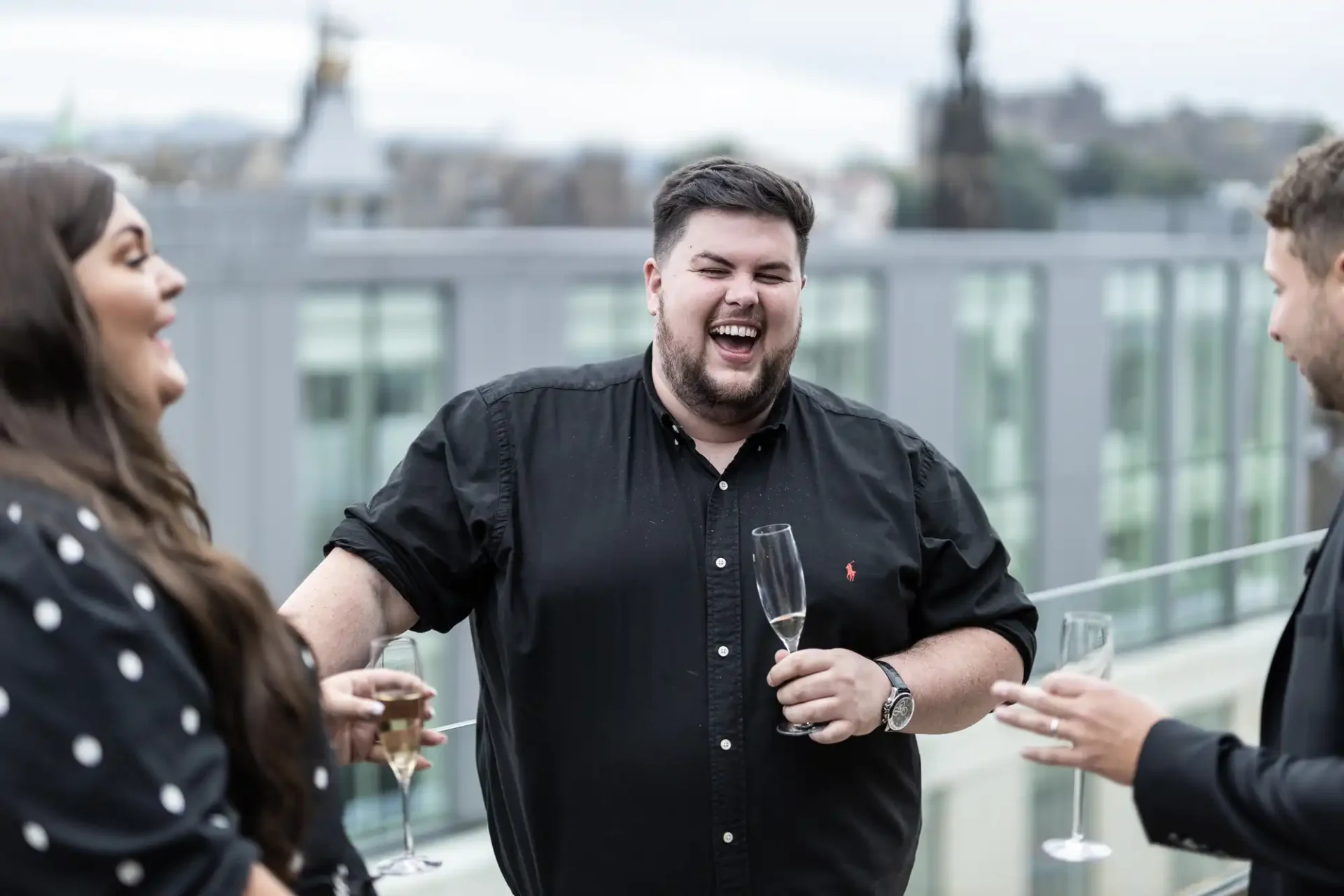 A man laughs joyously holding a glass of champagne, standing with friends on a rooftop, cityscape in the background.