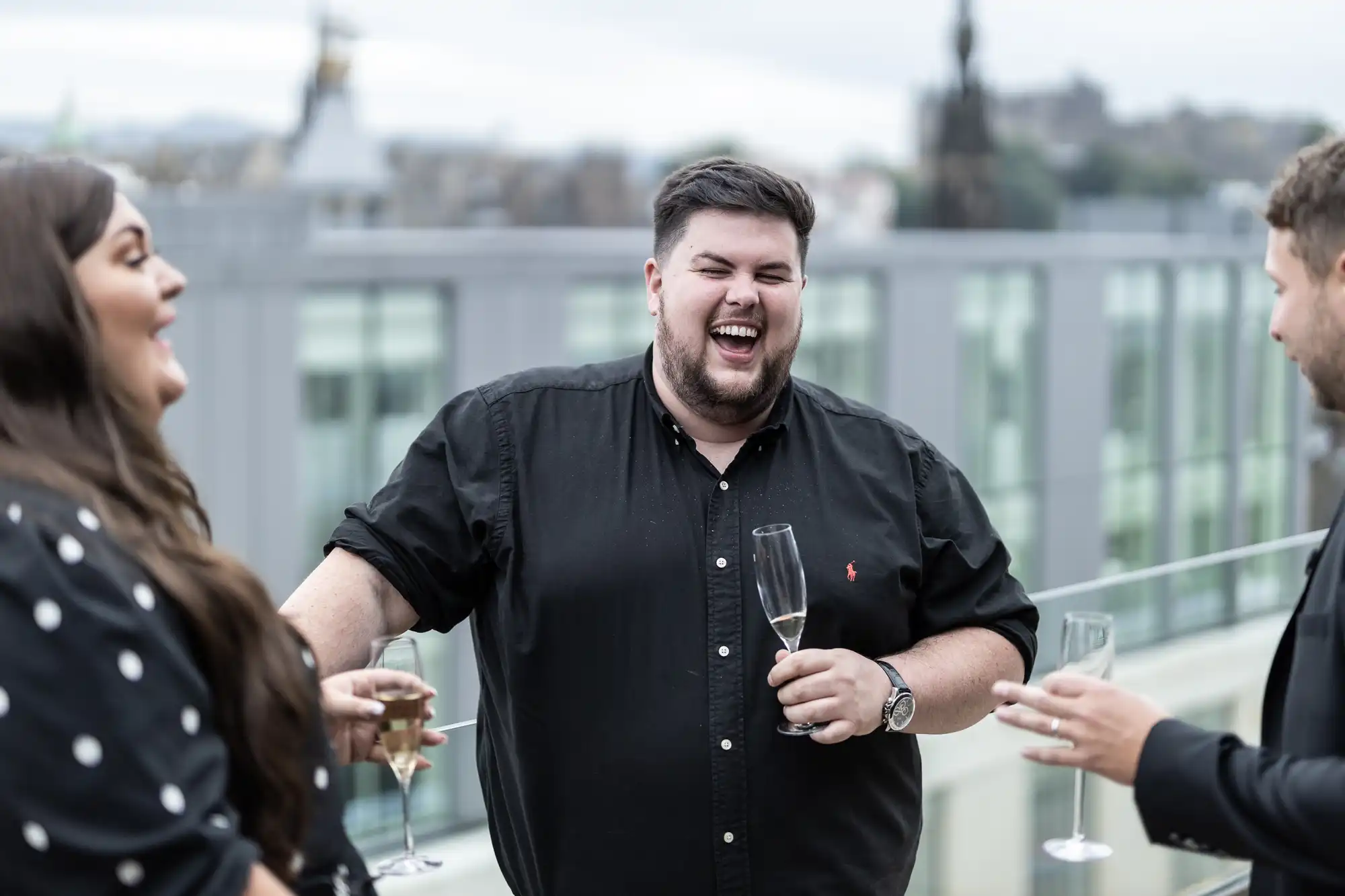 A man laughs joyously holding a glass of champagne, standing with friends on a rooftop, cityscape in the background.