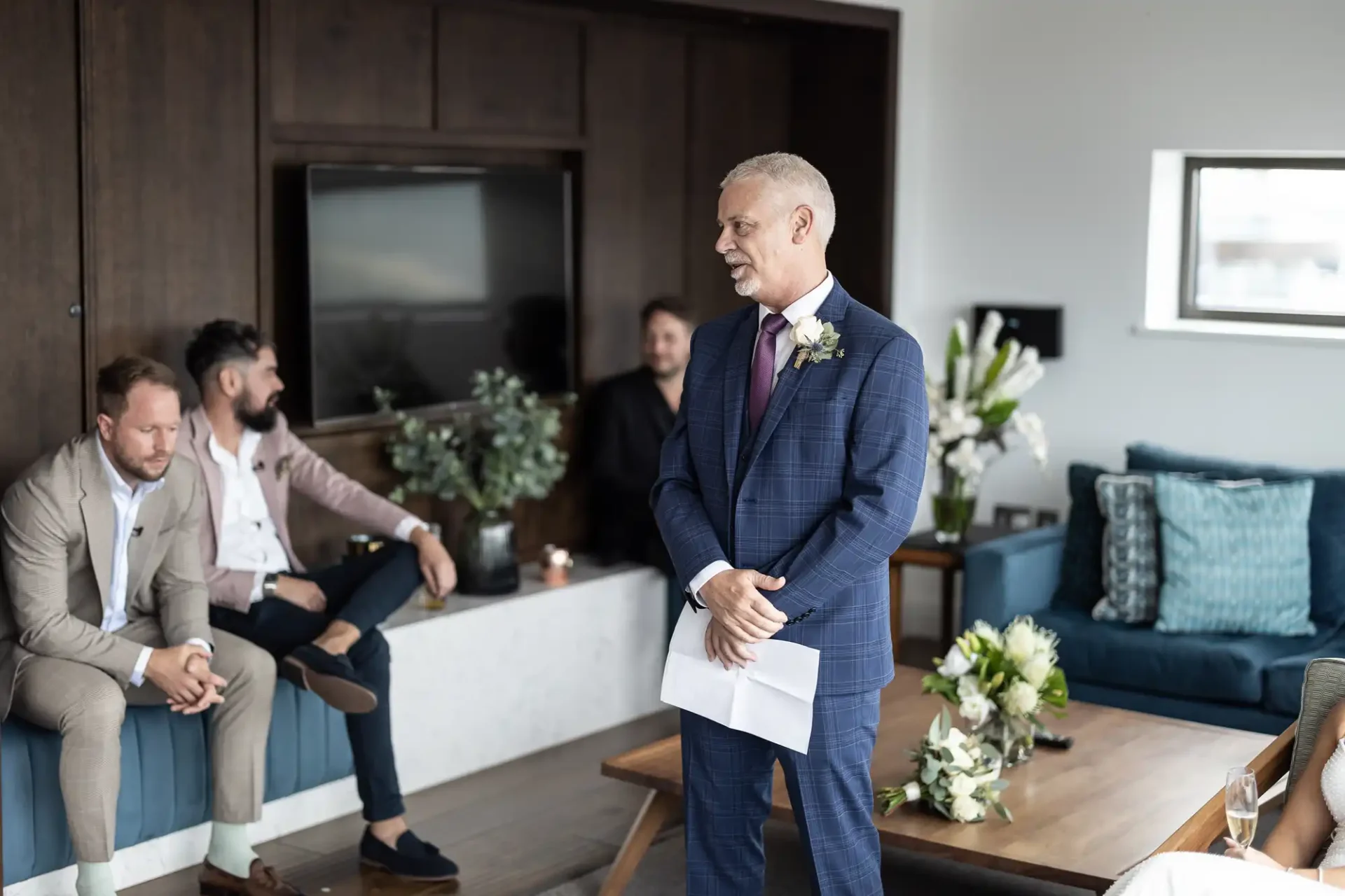 A man in a suit stands with a speech in hand at a wedding, while three guests listen attentively in a cozy living room.