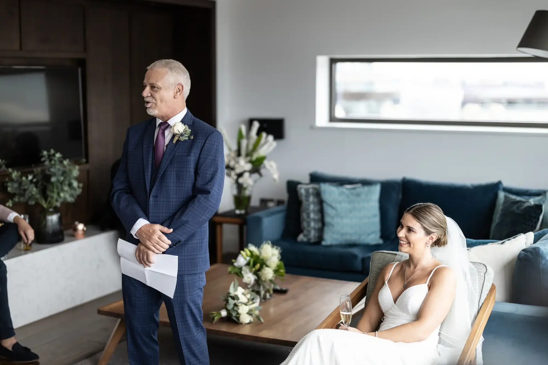 Father of the bride giving a speech with the bride seated and smiling in a modern living room setting.