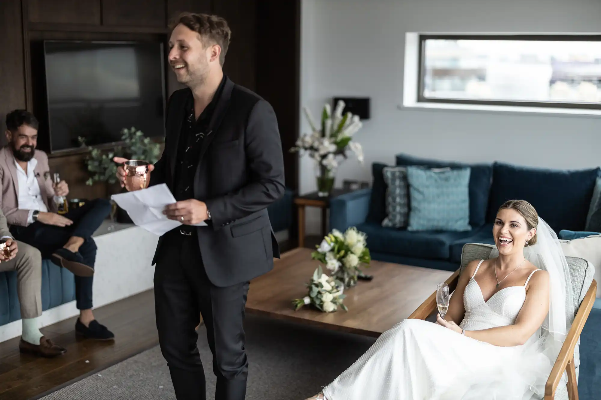 A man in a suit reading from a paper, smiling at a seated bride laughing joyously, with another guest smiling in the background in a modern living room.