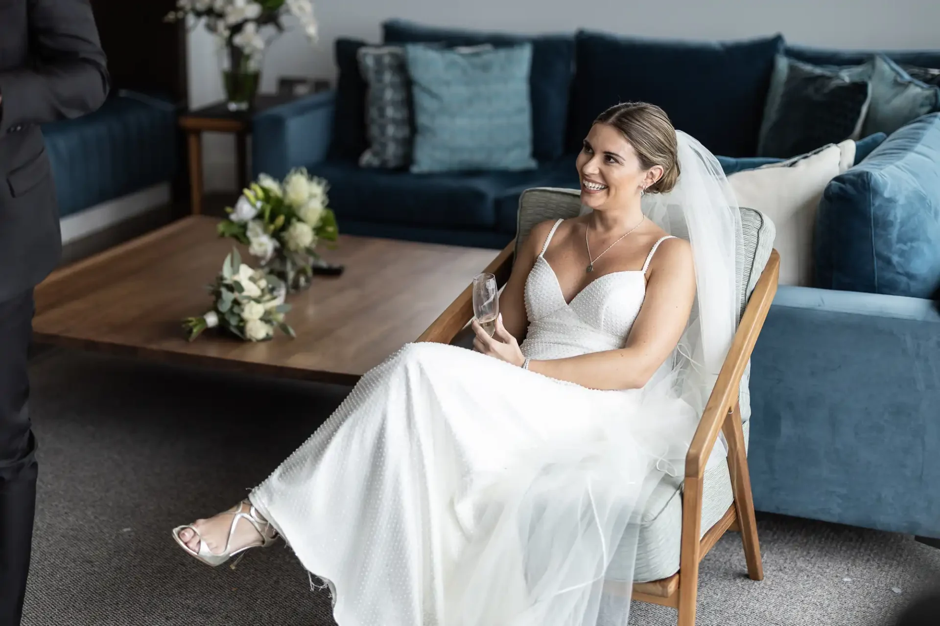 A bride in a white dress smiling, seated on a chair in a modern living room, holding a glass, with a man's torso visible on the left.