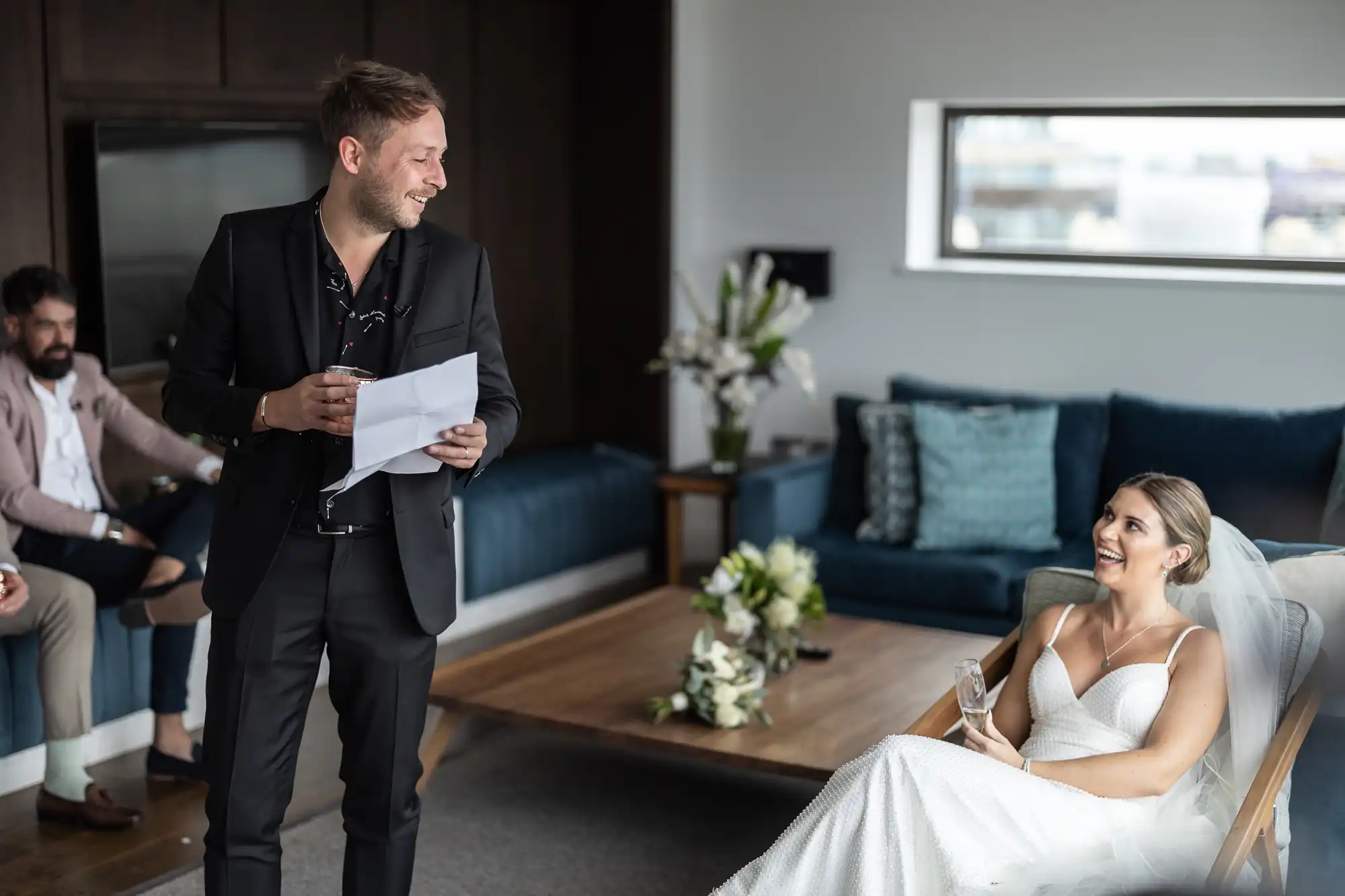 Man in suit smiling and holding papers stands near seated woman in white dress, with another man in background, in a modern room.