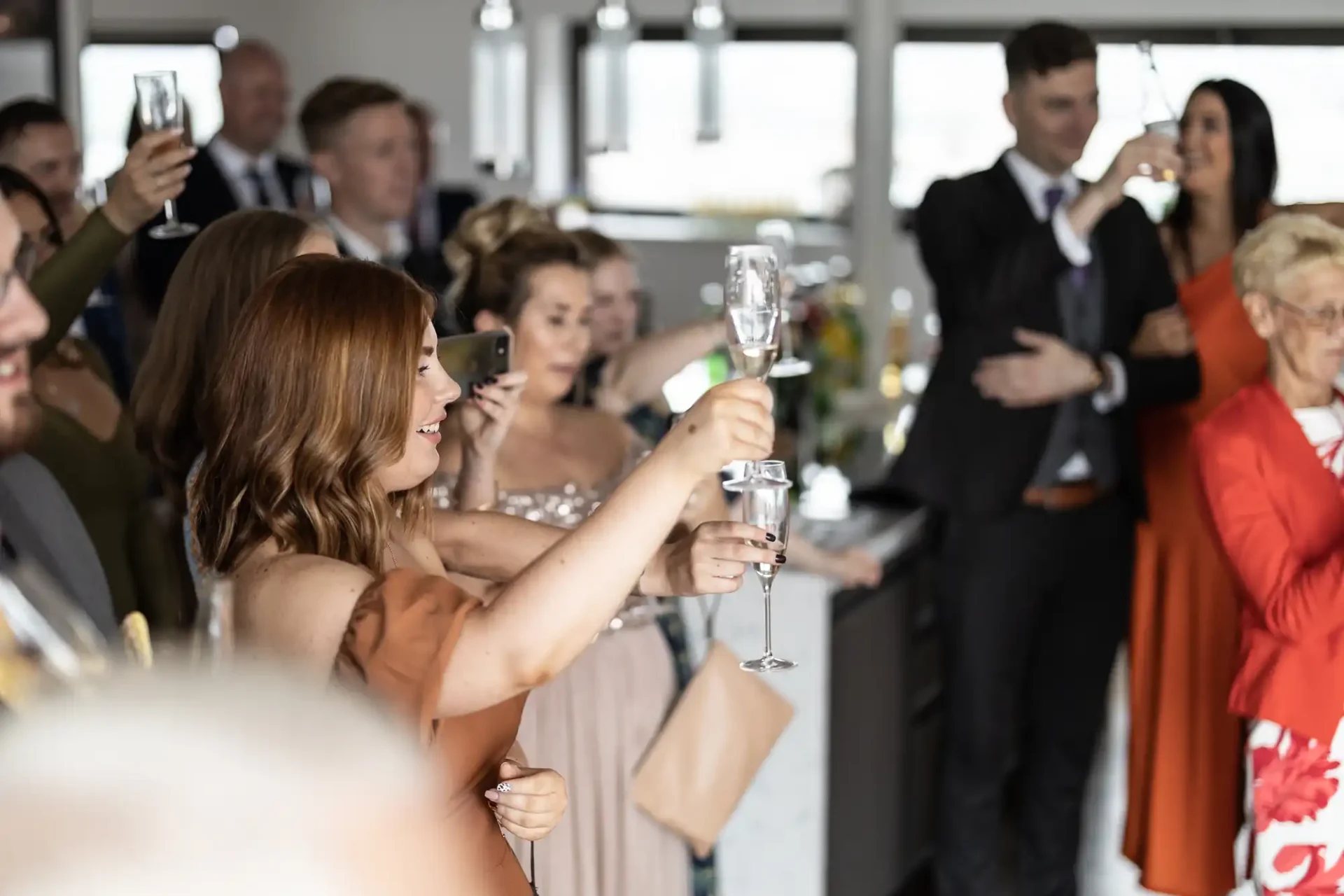 Guests holding up champagne flutes in a toast at a formal indoor gathering, focused on a woman with a raised glass in the foreground.