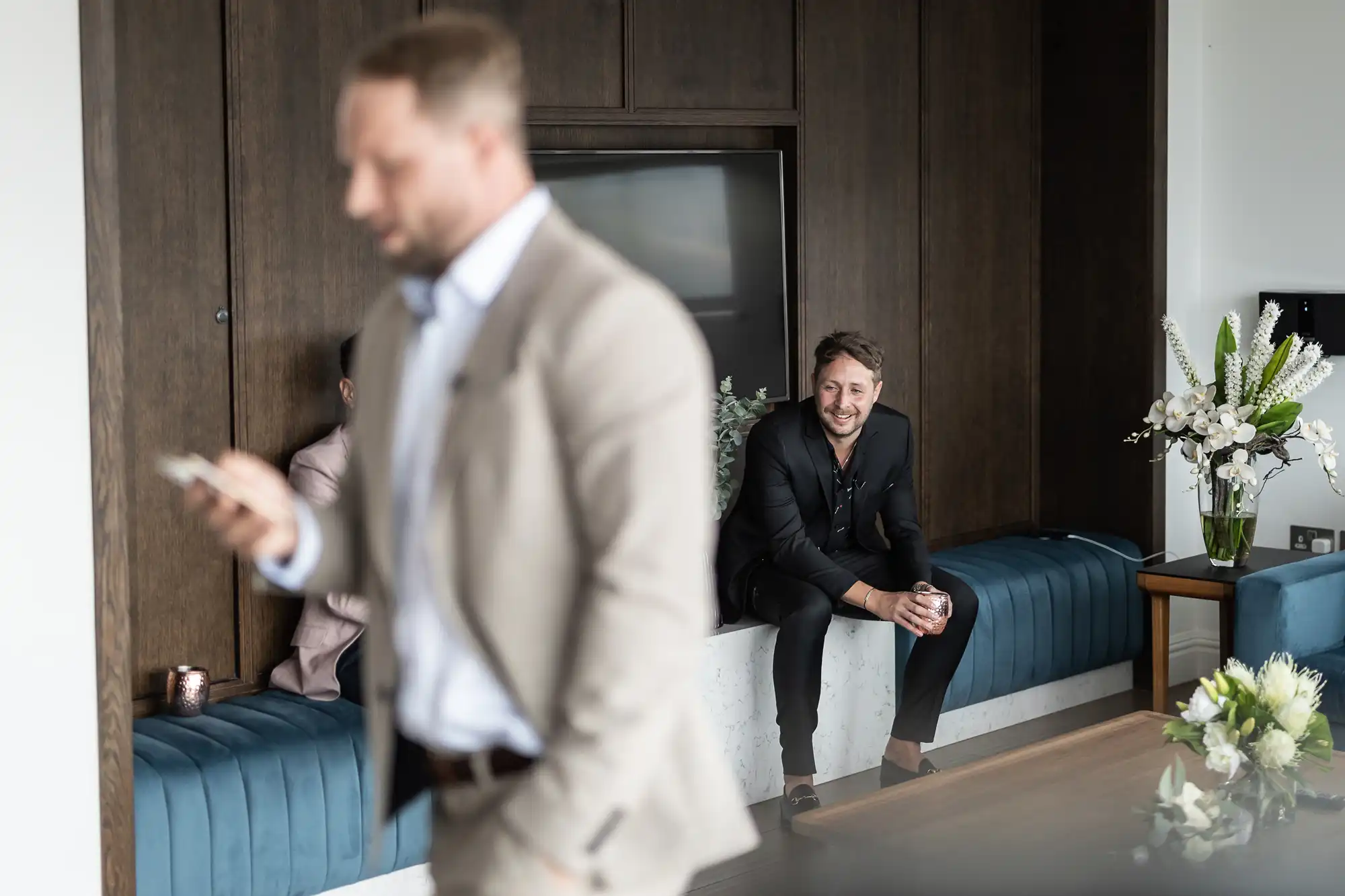 A blurry man in the foreground checking his phone with another man smiling seated in the background inside a modern room.