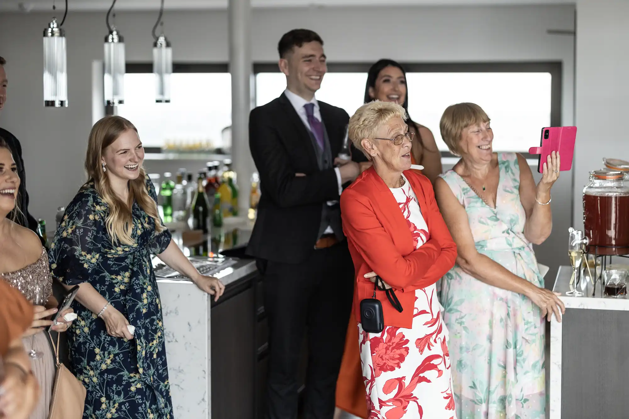 A group of people at a social gathering, smiling and watching something off-camera; one woman takes a photo with a tablet.