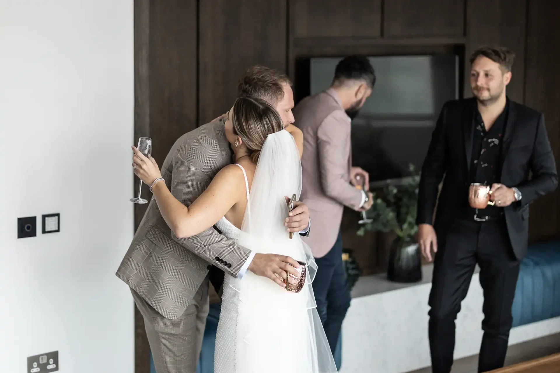 A bride hugs a guest at a wedding reception, holding a champagne glass, as two other guests converse in the background.