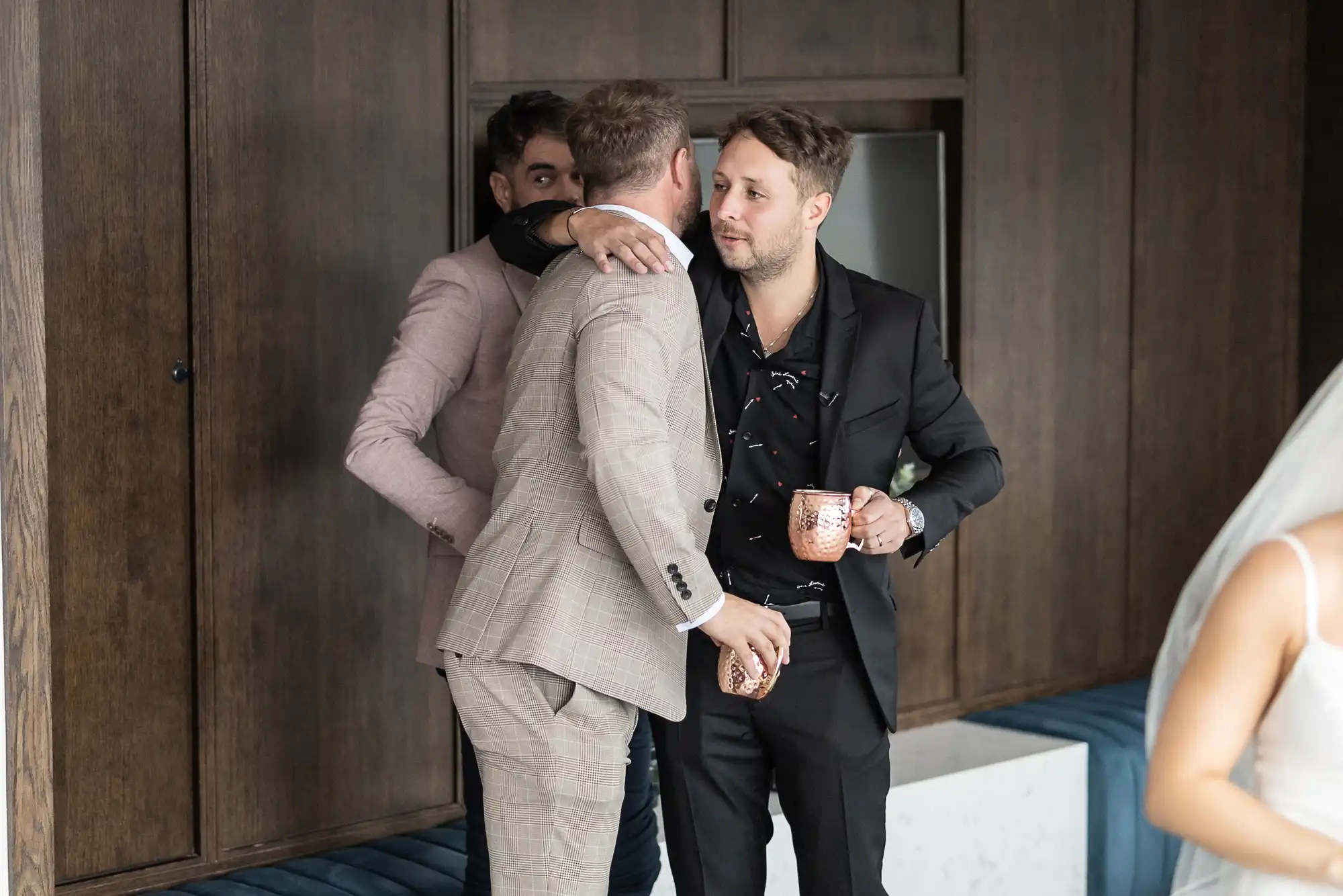 Two men in suits embrace warmly at a social gathering, one holding a drink, with others in the background.