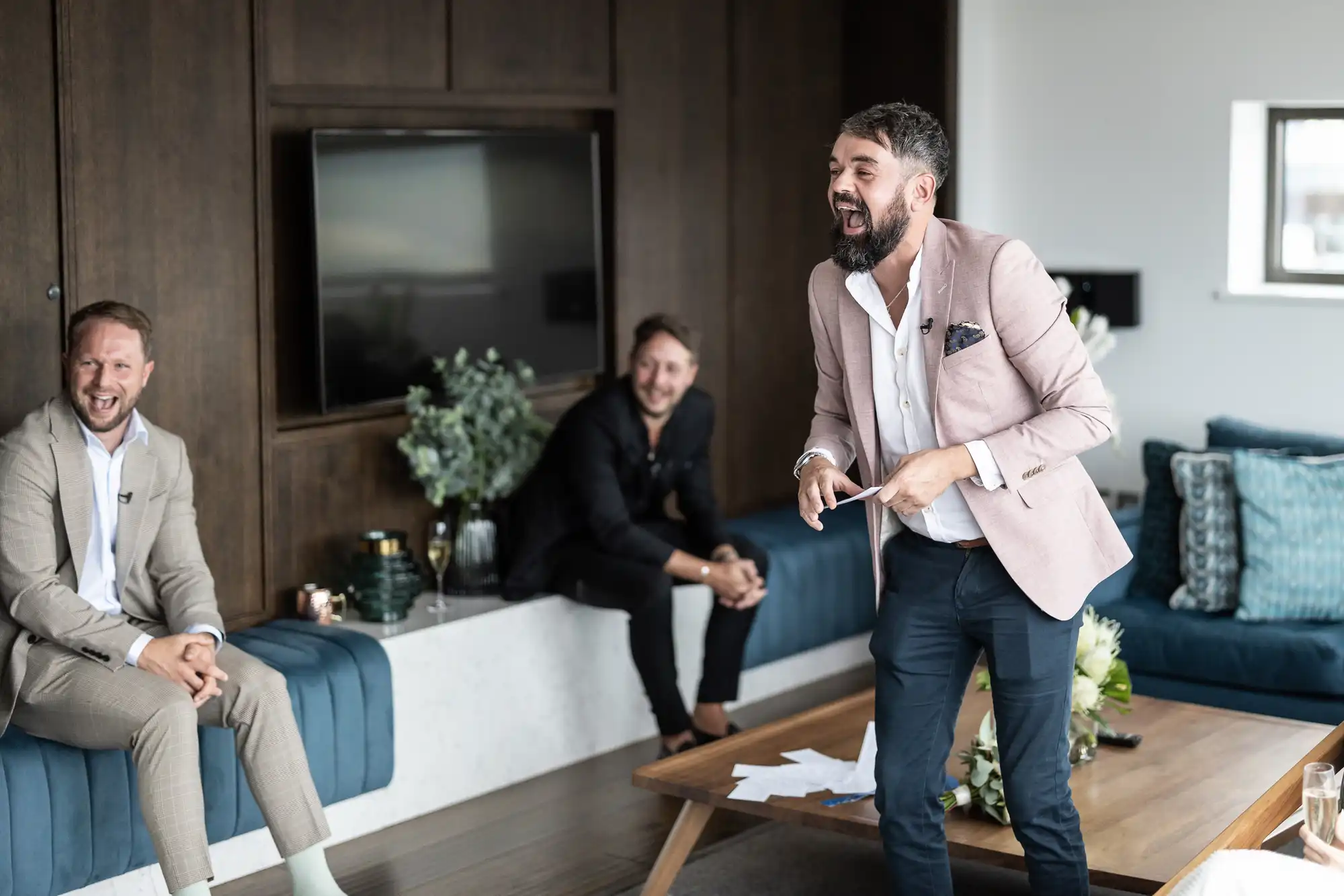 A man in a beige blazer passionately presenting in a modern living room setting with two laughing men seated behind him.