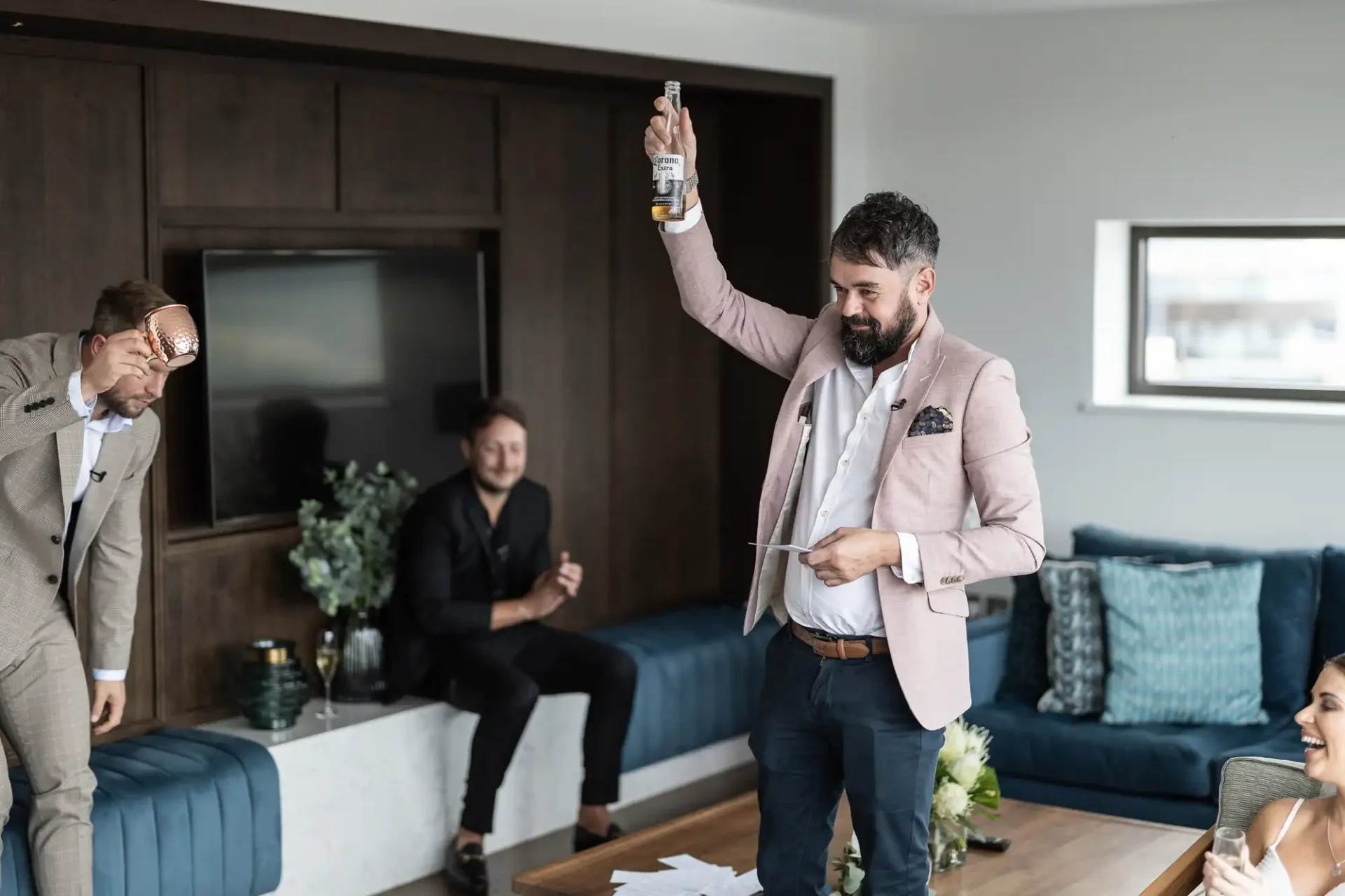 A man in a pink blazer raising a bottle in a toast while holding a speech, with three people watching in a modern living room.