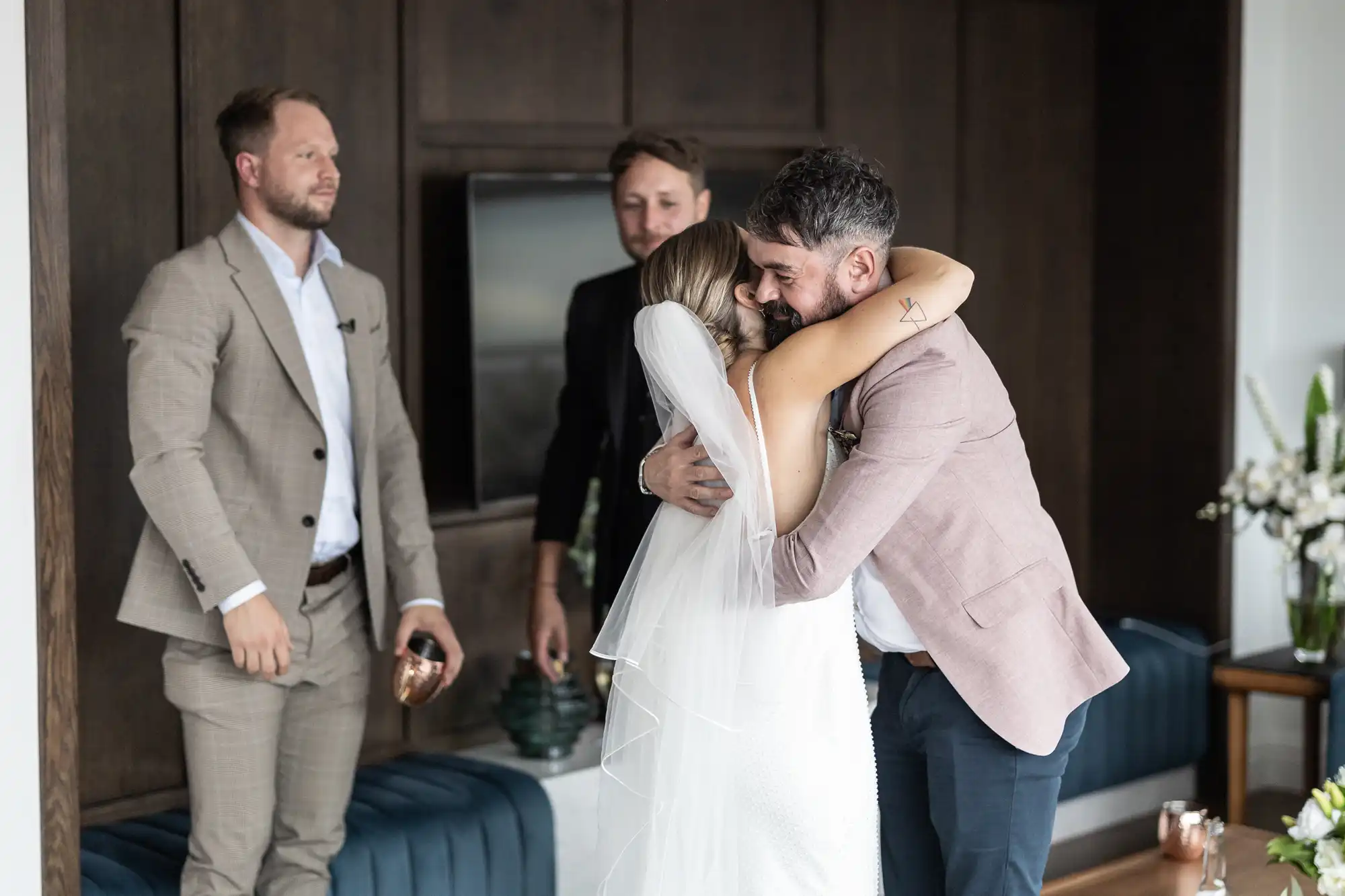 A bride and groom embracing warmly, watched by a smiling man in a suit, in a room with modern decor.