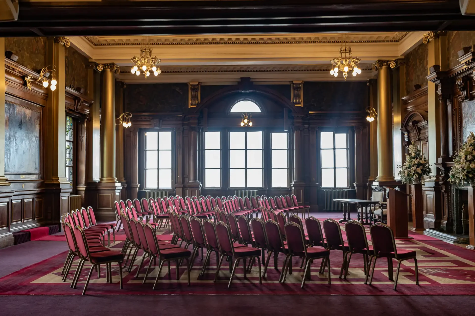 An empty, ornate room with rows of red chairs facing a podium, large windows, chandeliers, and decorative columns.