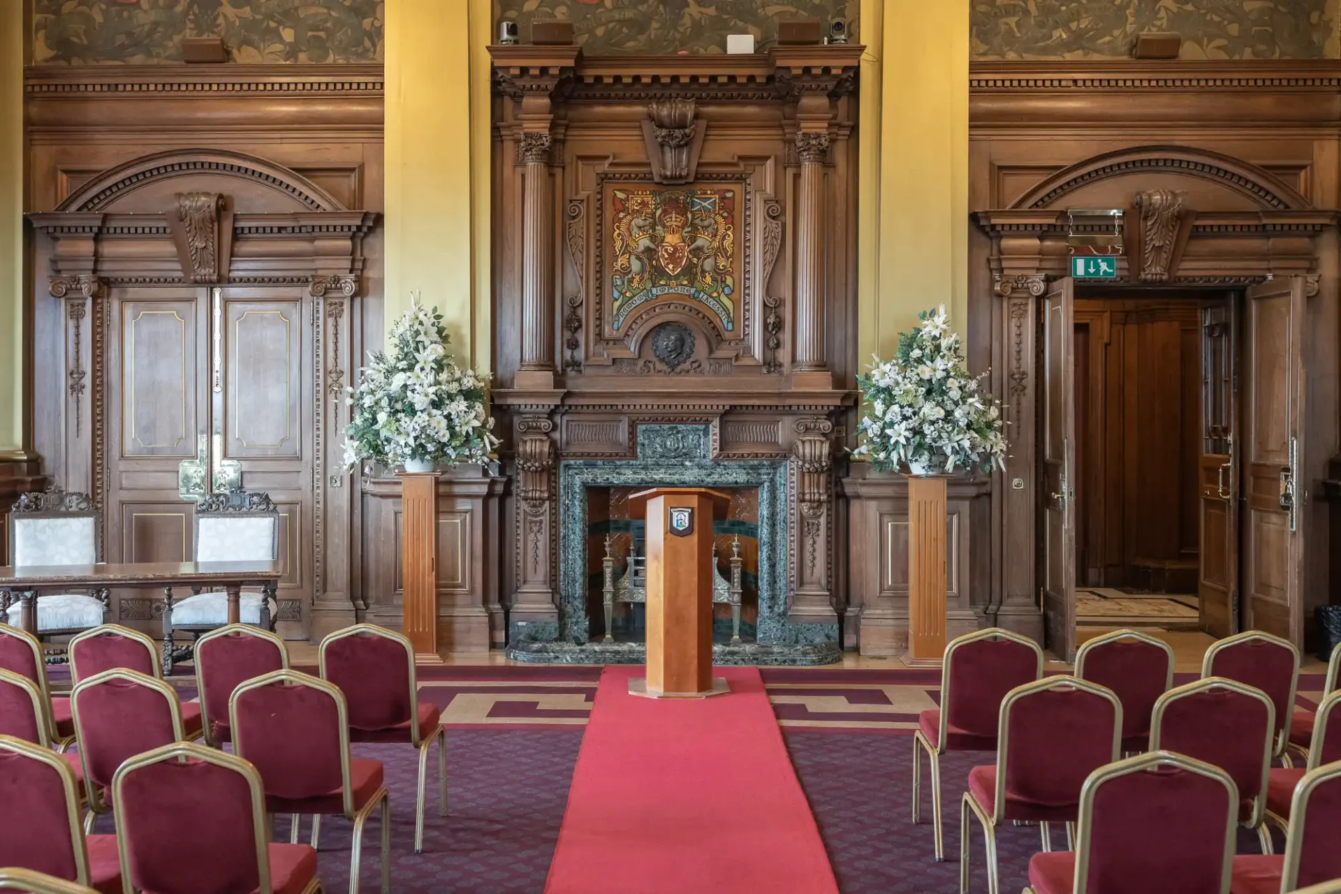 An ornate room with a red carpet aisle, wooden podium, and rows of red chairs. Elaborate wooden paneling and floral arrangements are present.