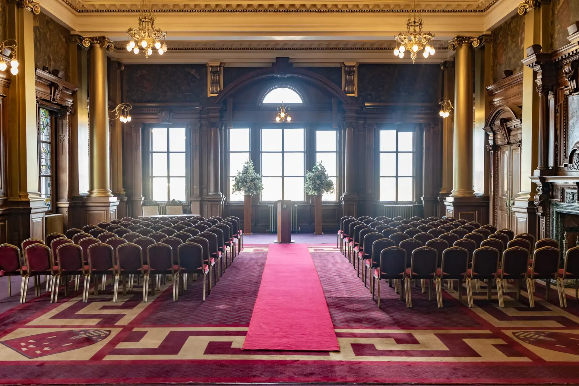 Ornate room set up for a ceremony with rows of chairs, a red carpet aisle, and a podium in front of large windows.