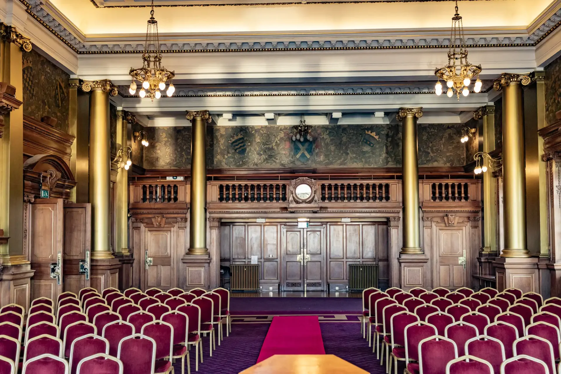 Ornate interior of a hall with red chairs, wooden panels, large columns, chandeliers, and a central aisle leading to a door and clock.