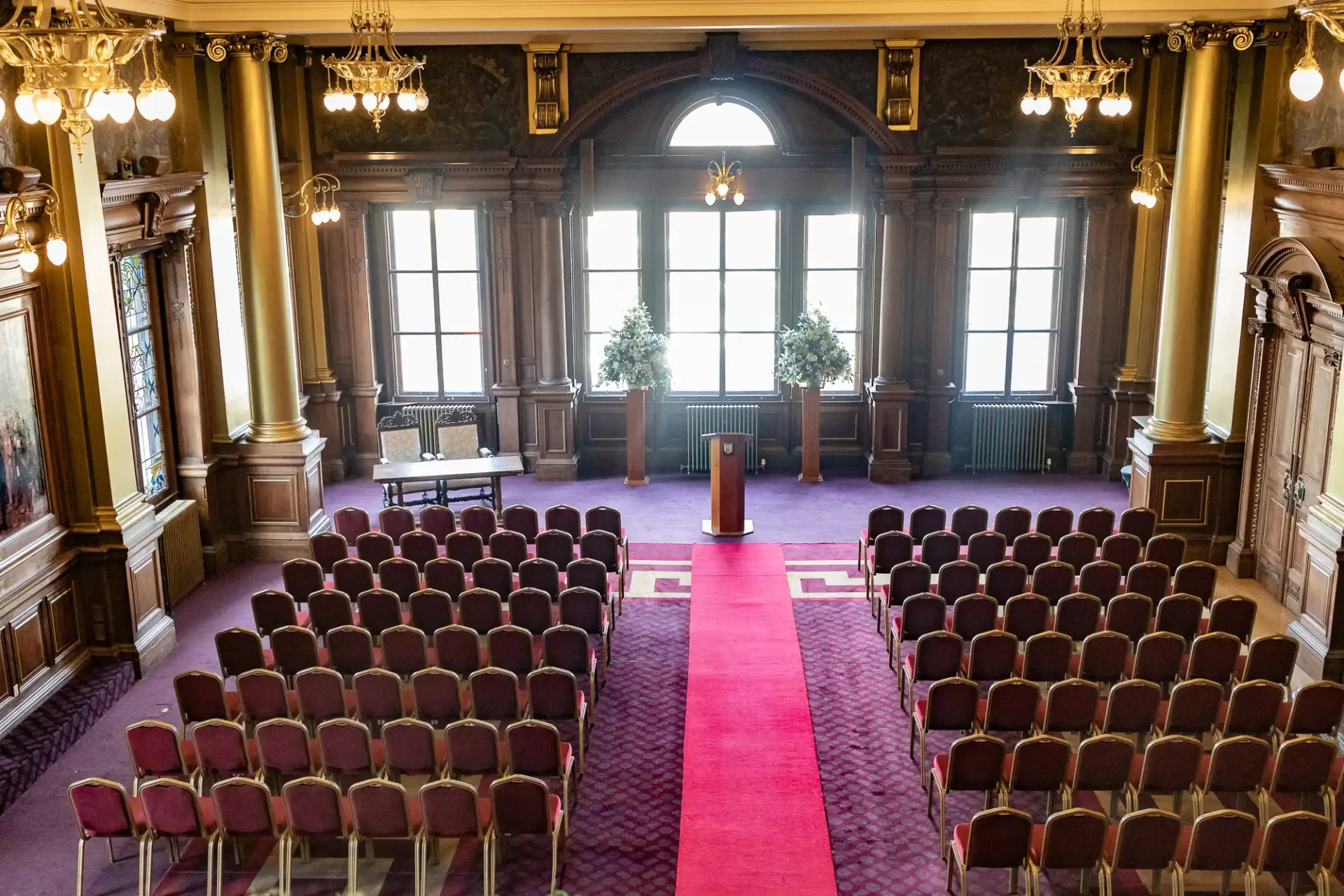 Ornate hall with rows of chairs facing a podium on a red carpet, large windows, and decorative columns.