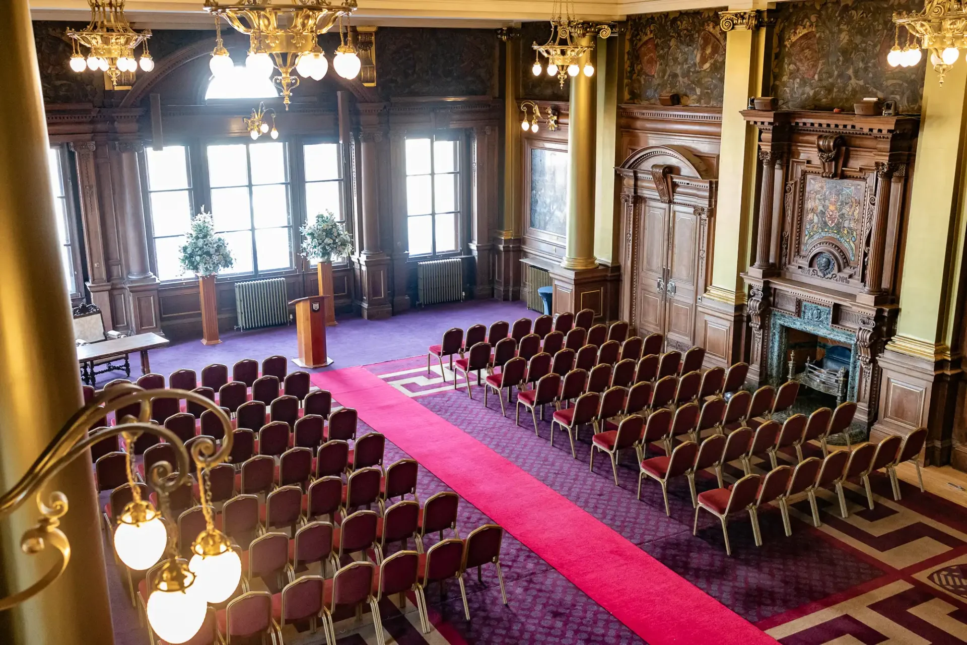 An ornate room set up for an event with rows of red chairs facing a podium, surrounded by wooden paneling and large windows.