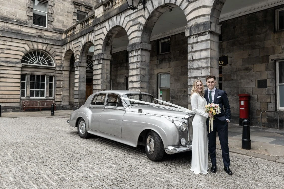Newlywed couple stand in front of a silver Bentley car in the Quadrant