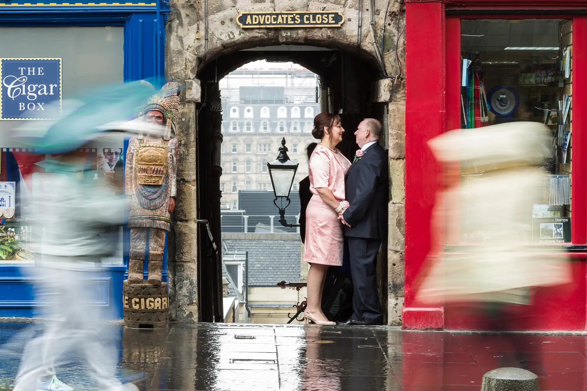 A couple in formal attire stands close together under the "Advocate's Close" sign between a blue shop and a red shop, with blurred pedestrians passing by in the foreground.