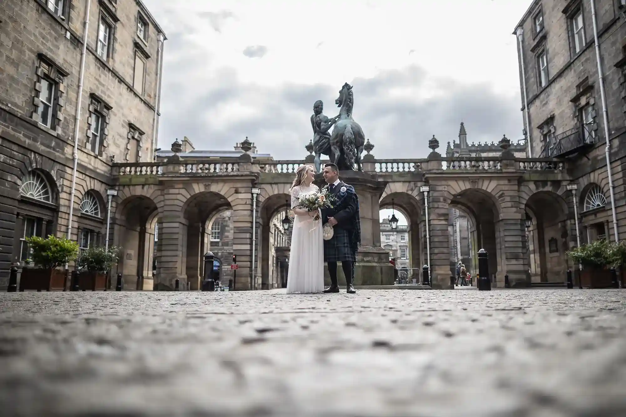 A couple in wedding attire stands in front of an equestrian statue, surrounded by a historic stone building with arches.