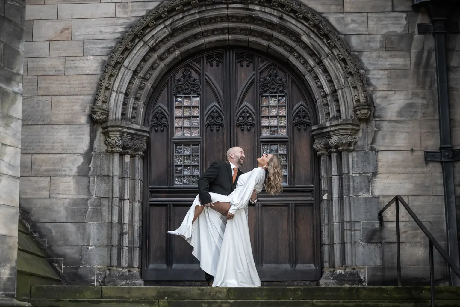 A couple stands on stone steps in front of a large, ornate wooden door. The man in a black suit dips the woman in a white dress, and both are smiling.