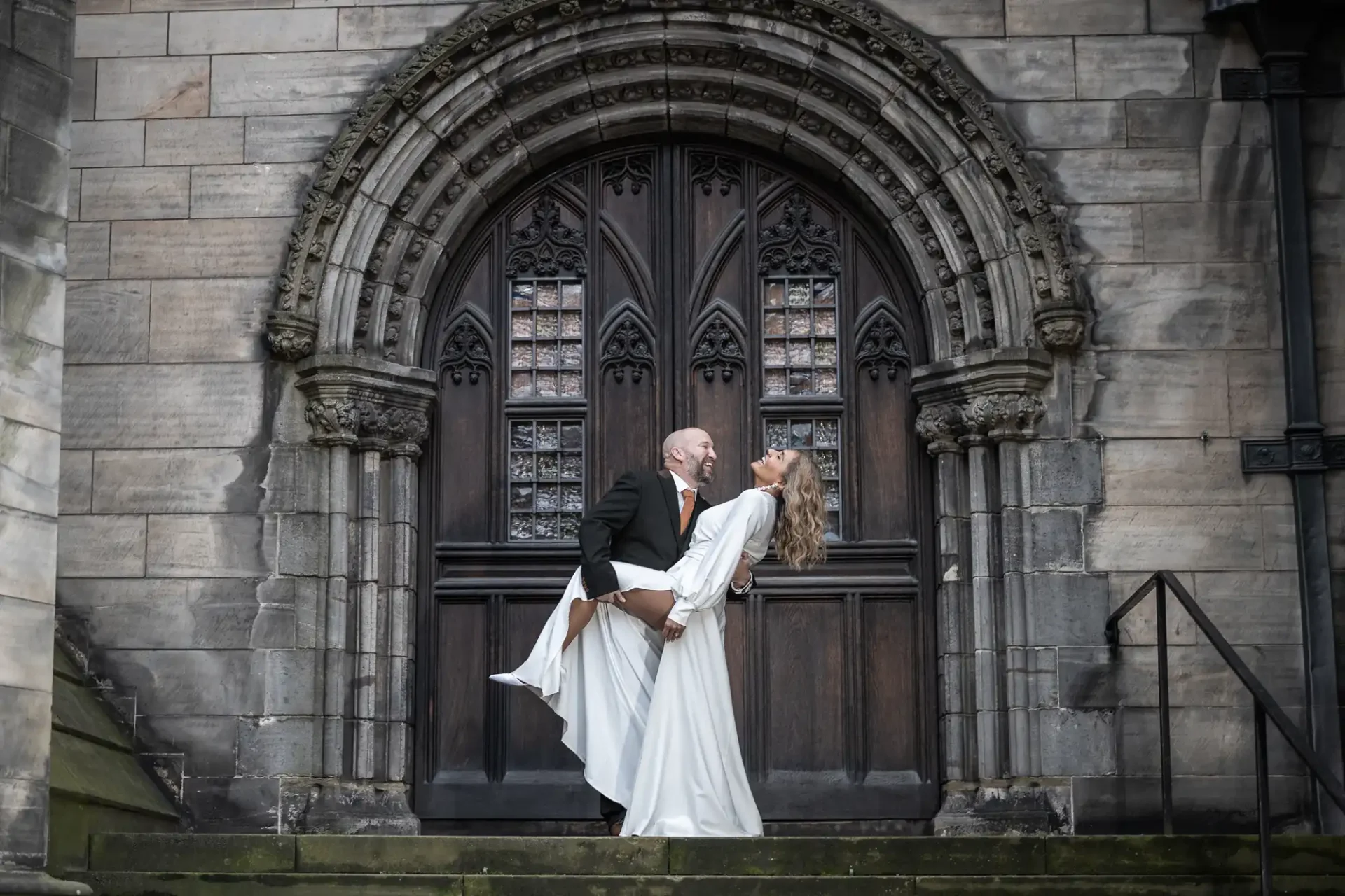 A couple stands on stone steps in front of a large, ornate wooden door. The man in a black suit dips the woman in a white dress, and both are smiling.