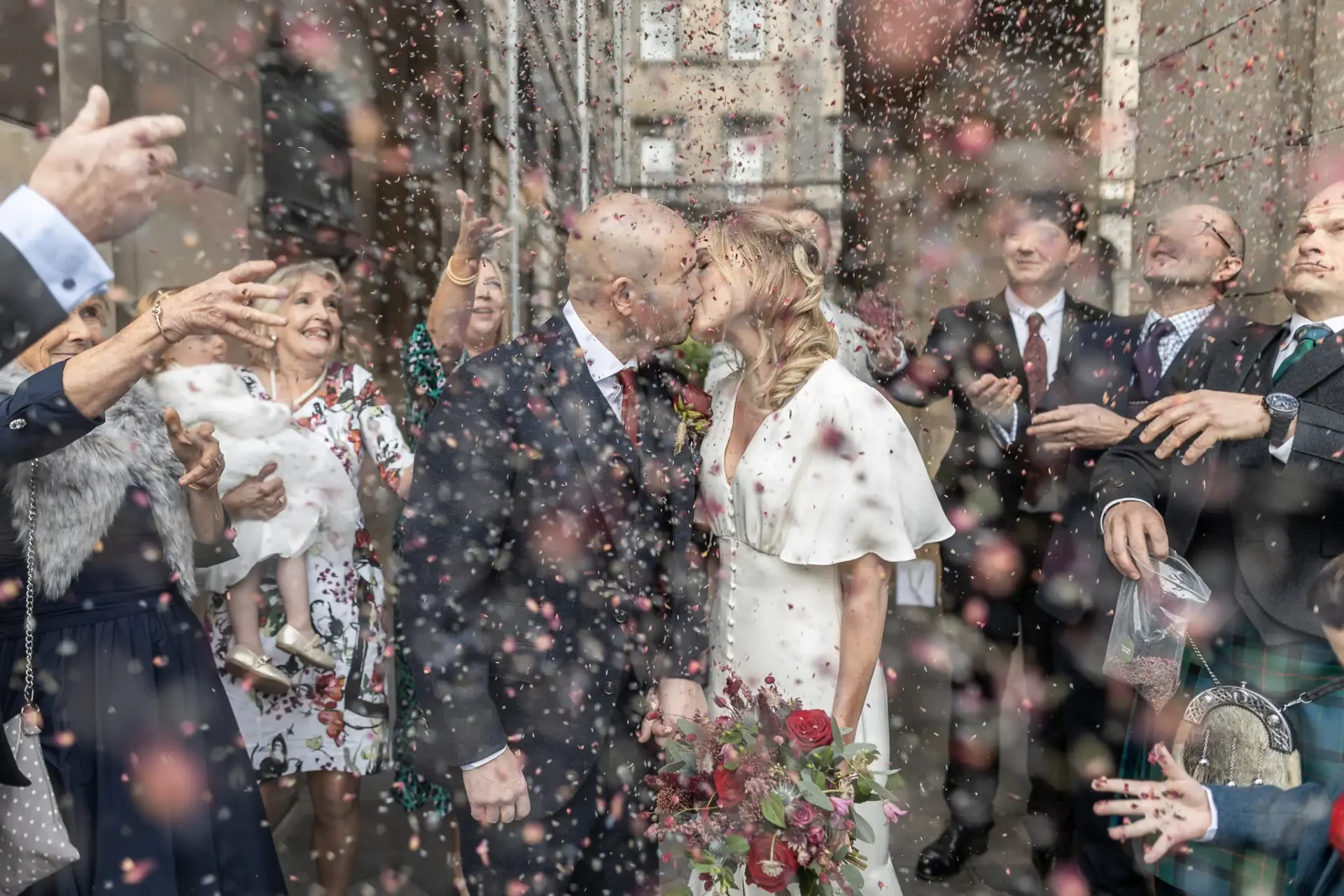 A bride and groom kiss surrounded by guests throwing confetti. The bride holds a bouquet, and guests are smiling, celebrating their wedding.