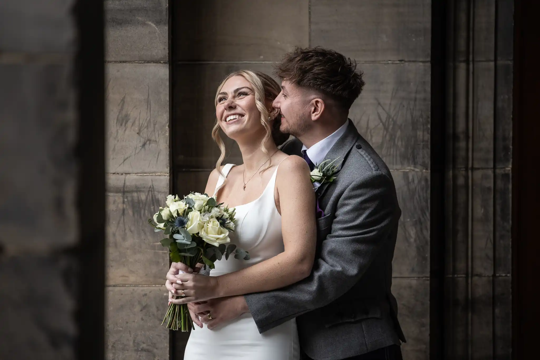 City Chambers wedding couple embraces in a dimly lit hallway, with the woman holding a bouquet of white flowers. Both are dressed formally; the man wears a suit, and she is in a white dress, smiling, taken by Edinburgh wedding photographer.