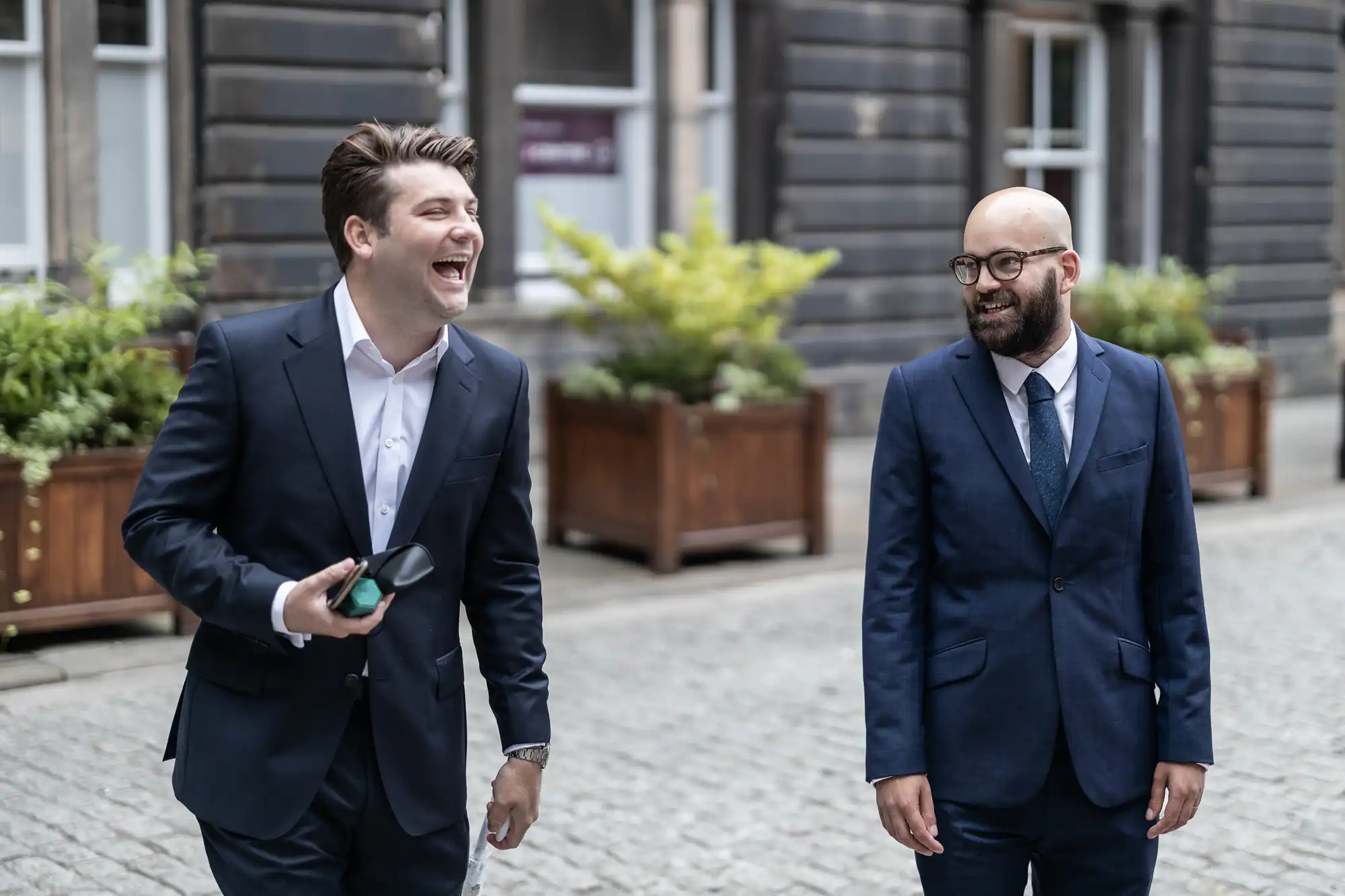 Two men in business suits walking outdoors, one laughing heartily and holding a smartphone, the other smiling and bald, in a quaint urban setting.