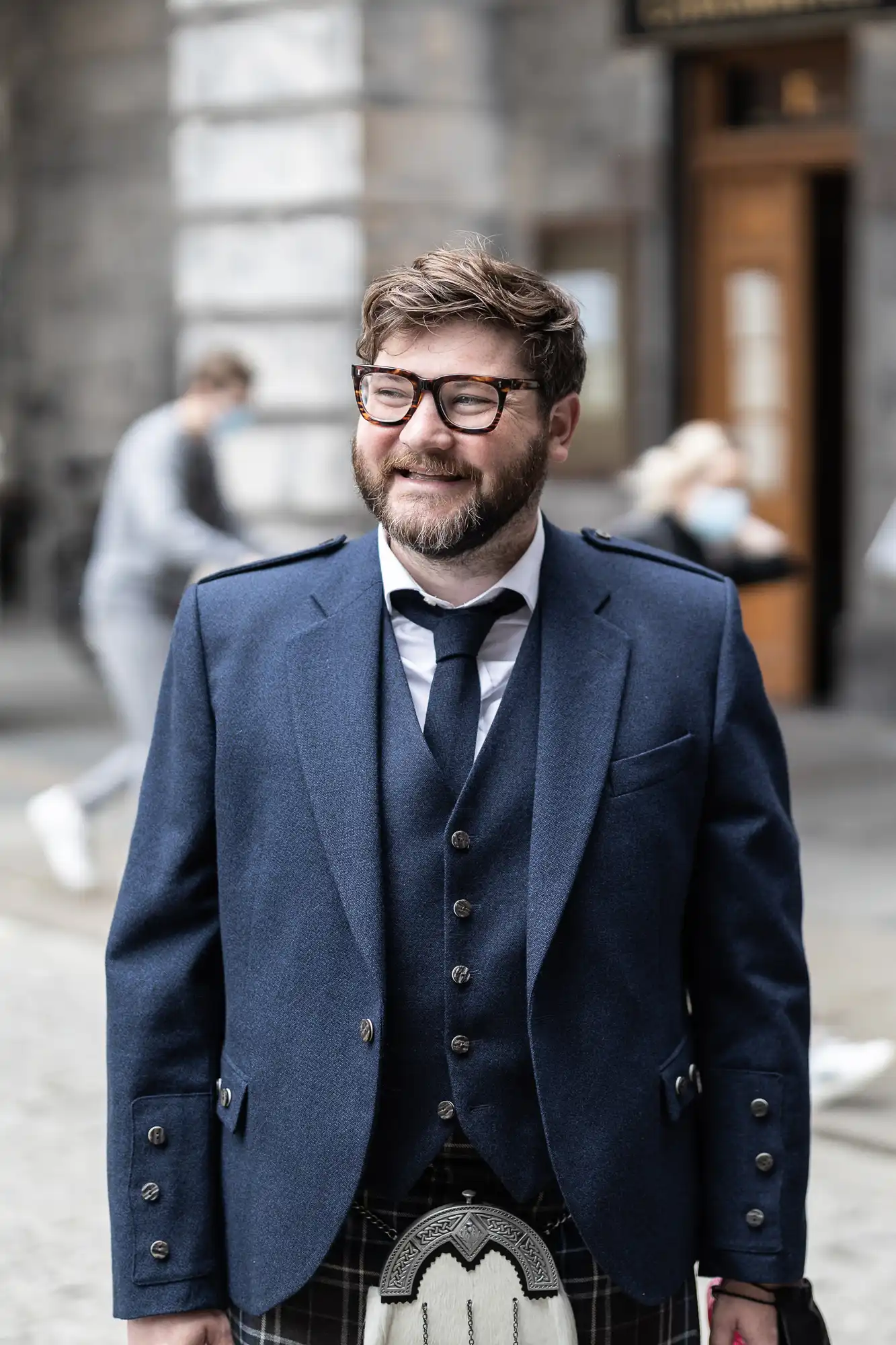 A man in a blue blazer and kilt smiling on a city street.