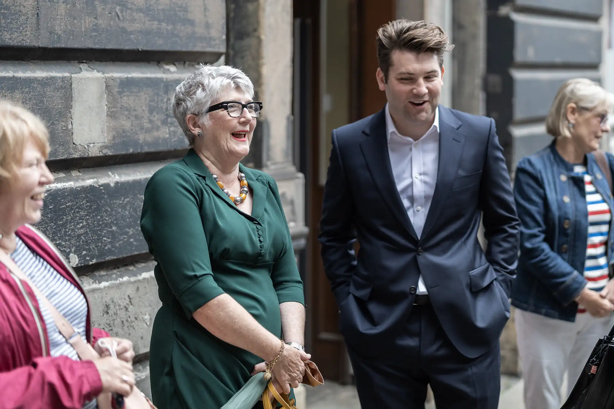 An older woman in a green dress laughs joyfully next to a young man in a suit, both standing on a city street.