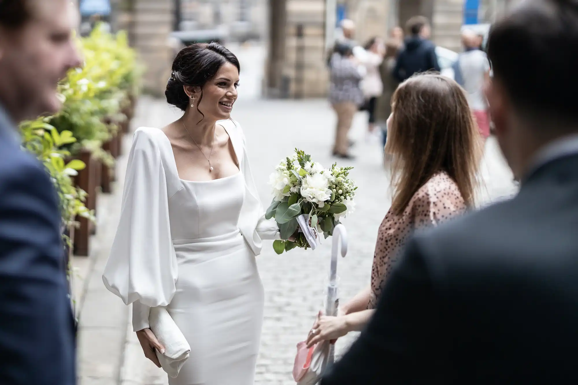 A bride with a bouquet, smiling as she walks through a crowd in a city setting, wearing a white dress with a cape.