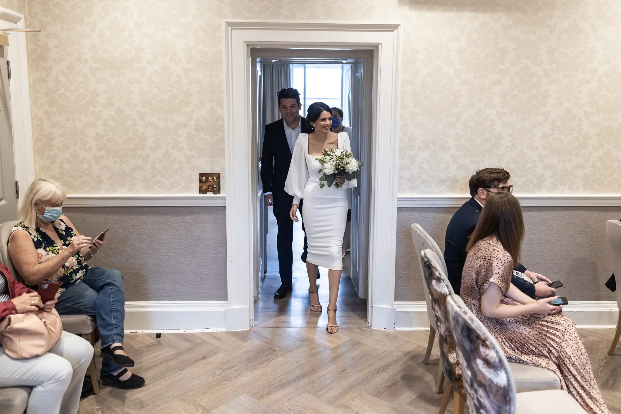 A bride and groom walking through a doorway smiling, while seated guests look at their phones, in an elegantly decorated room.