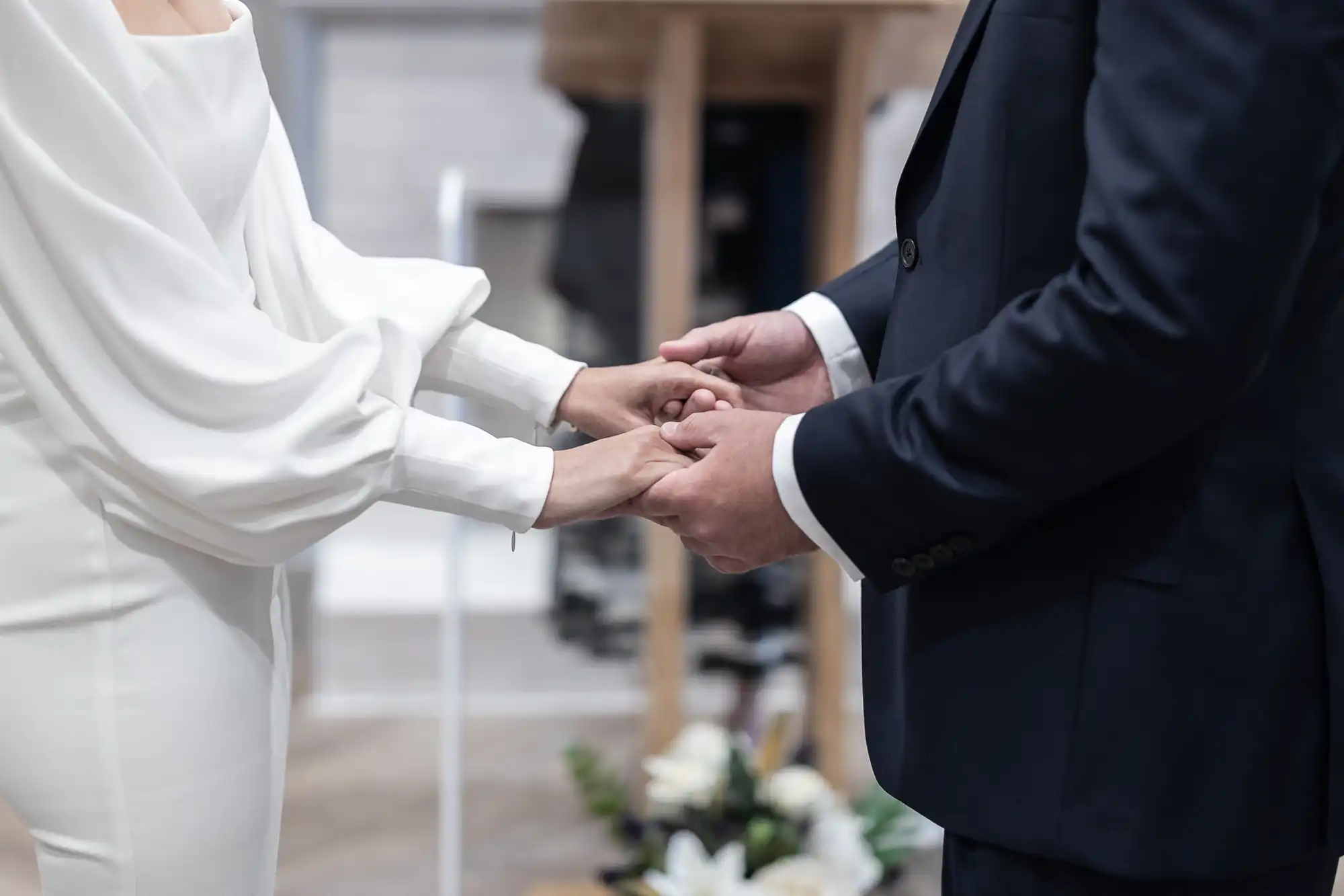 A bride and groom holding hands during a wedding ceremony, with a focus on their hands and blurred background.