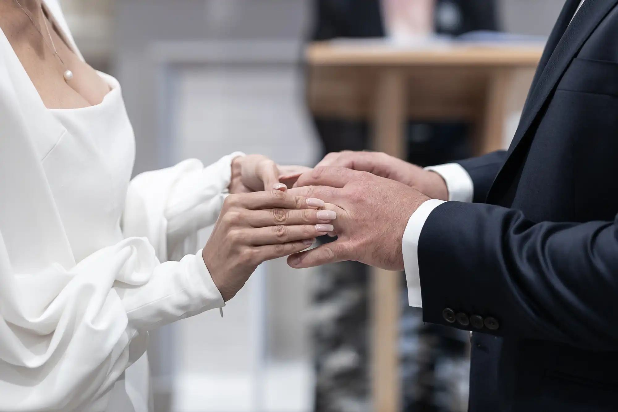 A bride and groom exchange wedding rings, focusing on their hands clasping over a blurred background.