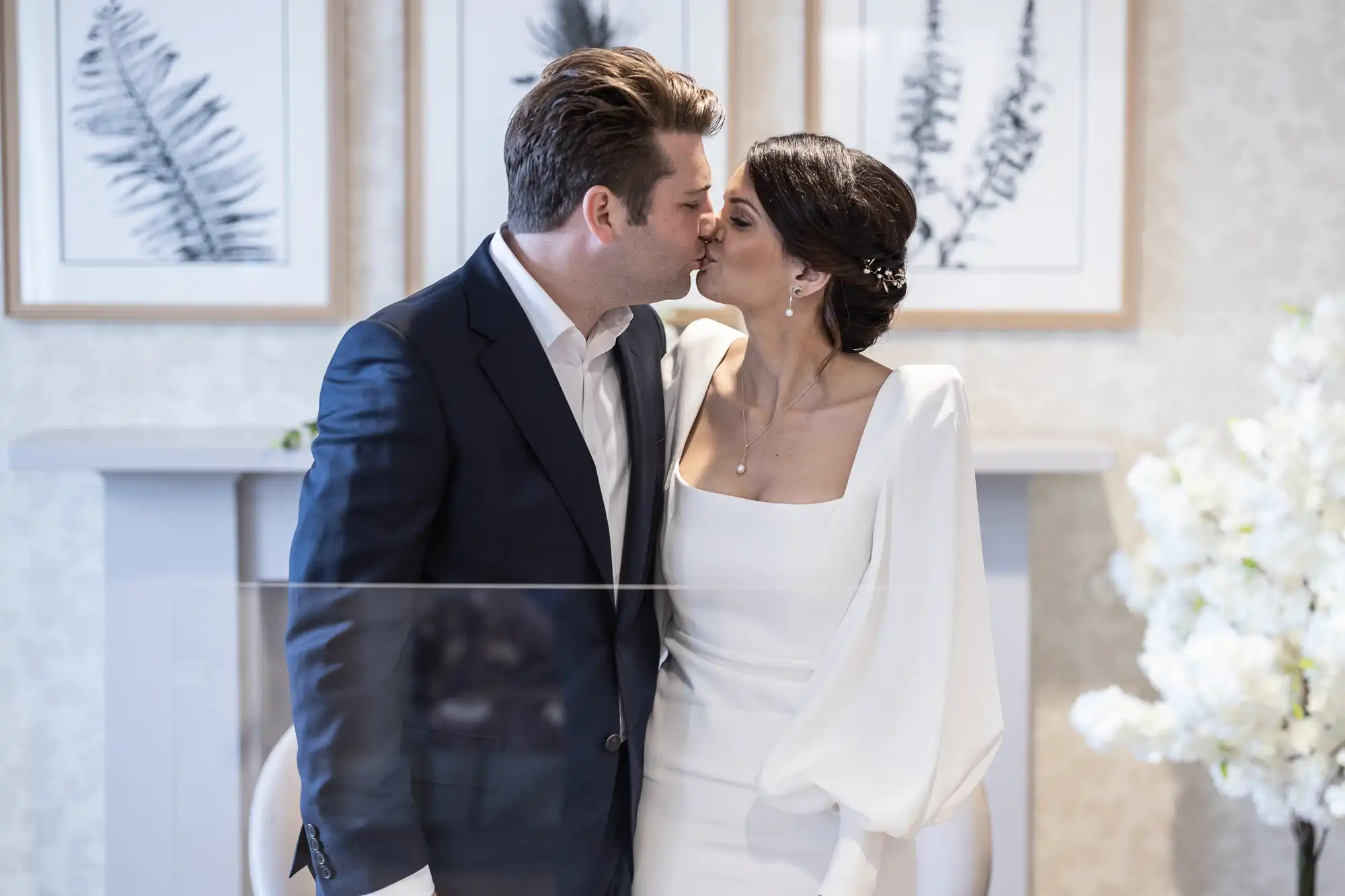 A bride in a white gown and a groom in a black suit kiss softly in a well-decorated room with white flowers and elegant art on the walls.