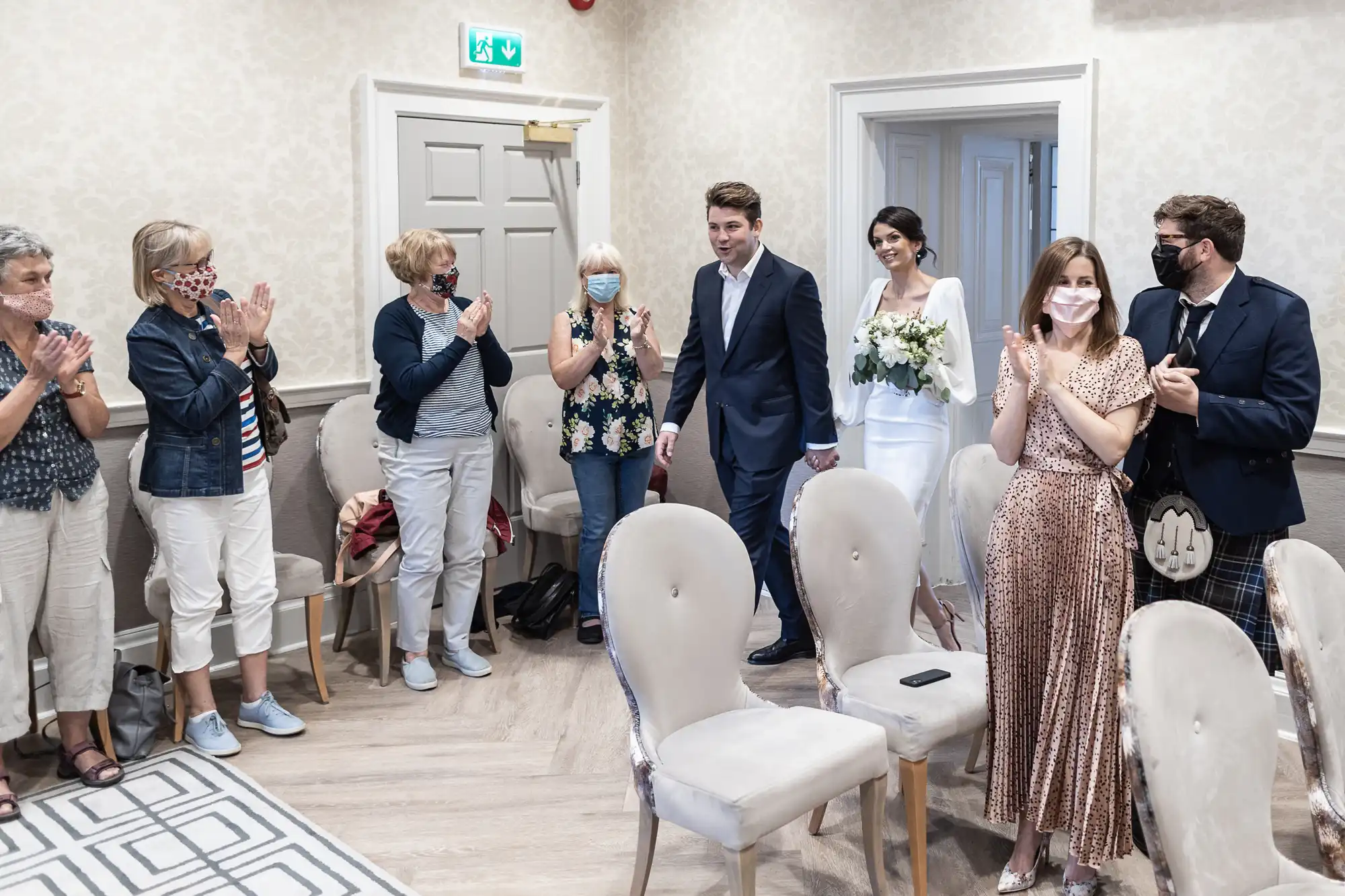 A newlywed couple walks through a small room receiving applause from guests wearing masks; the setting is casual and intimate.