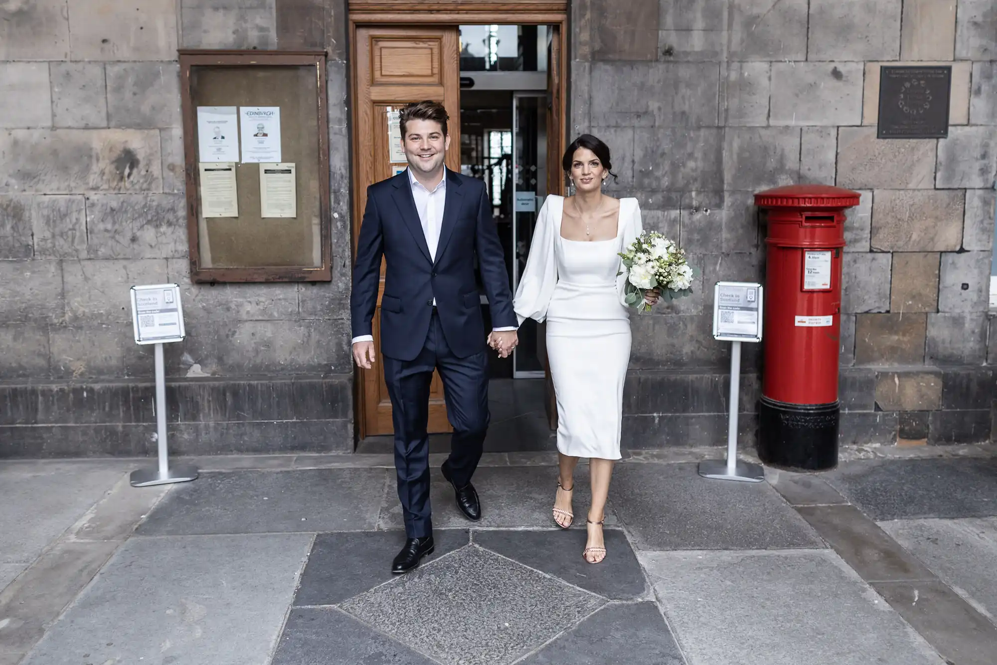 A newlywed couple holding hands and smiling, exiting a stone building, walking past a red post box. the bride wears a white dress and carries a bouquet.