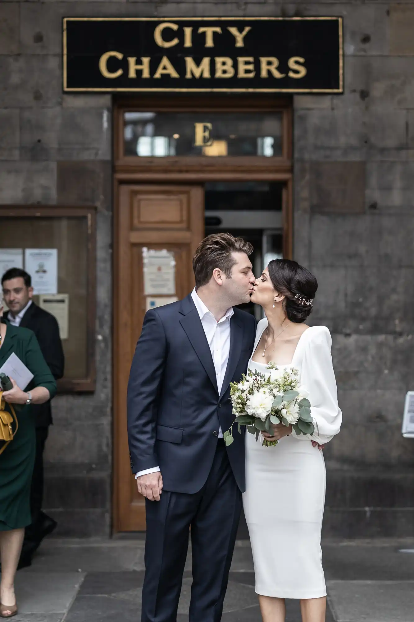A newlywed couple kissing outside the city chambers, with the bride holding a bouquet and the groom in a suit.