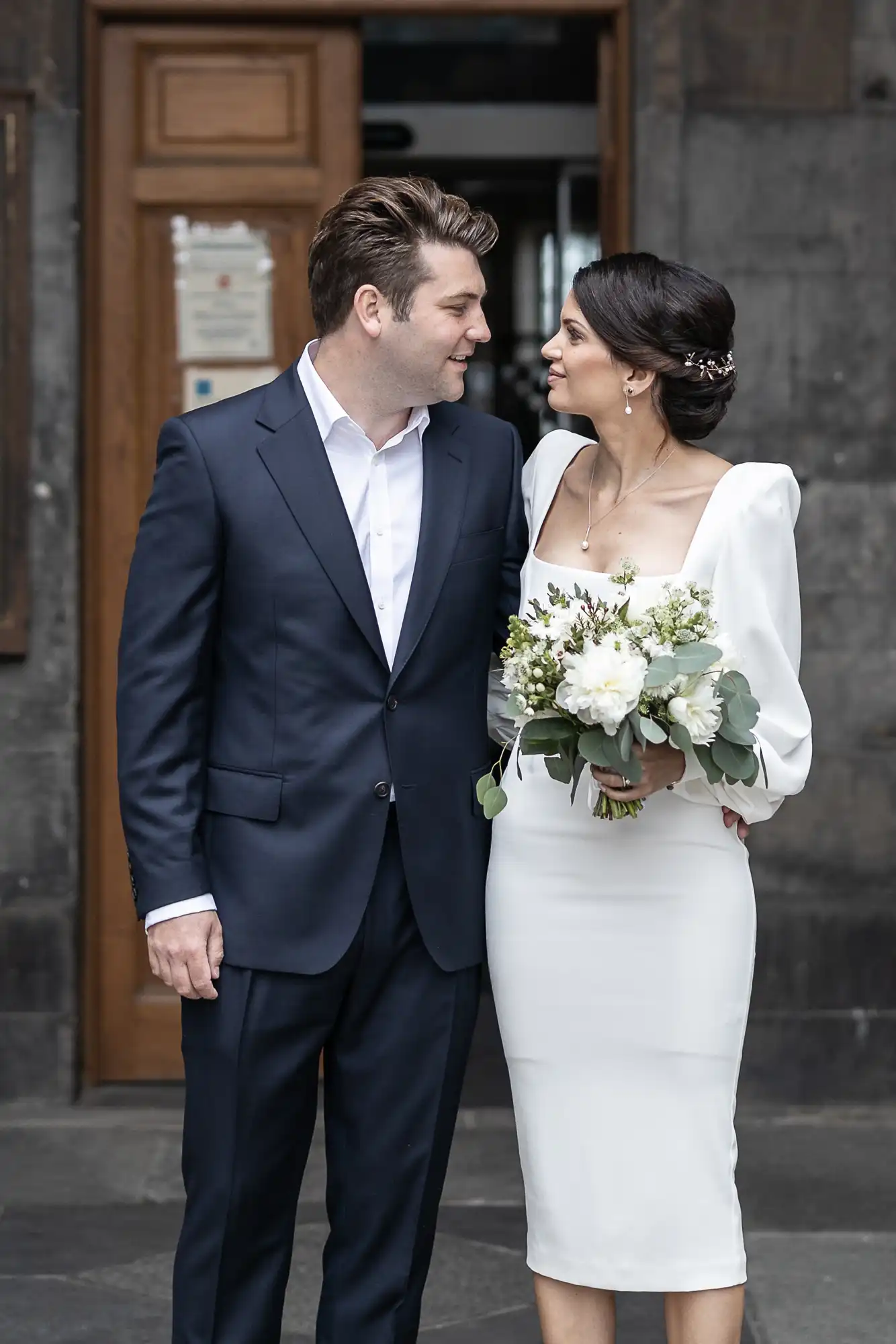 A bride in a white dress with a bouquet and a groom in a navy suit smiling at each other in front of a building with a wooden door.
