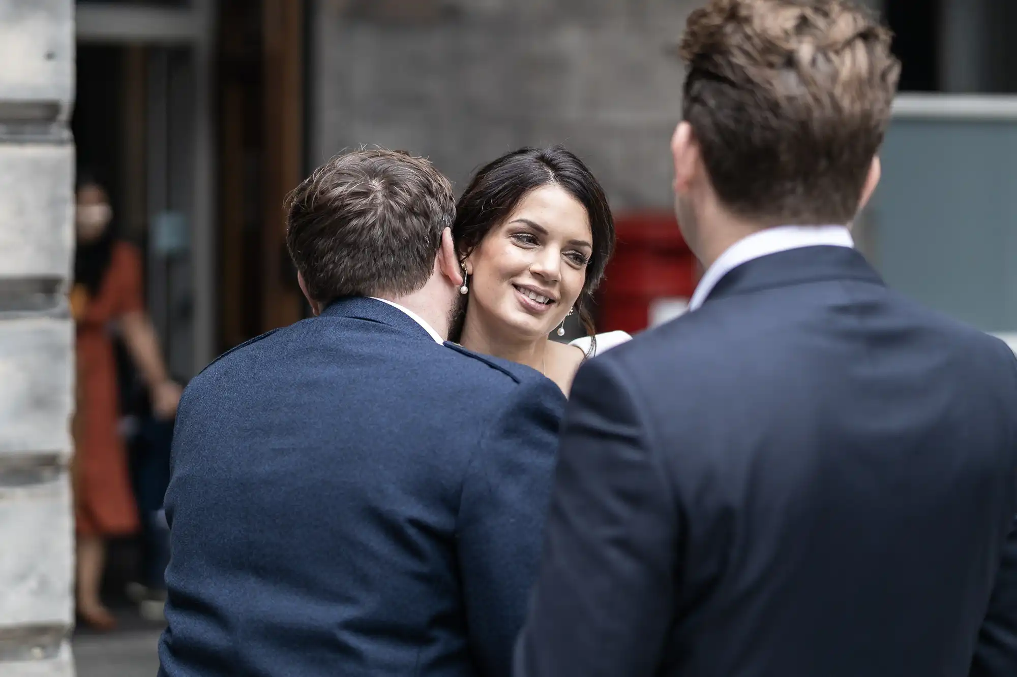A woman smiles while conversing with two men in suits at an outdoor gathering.
