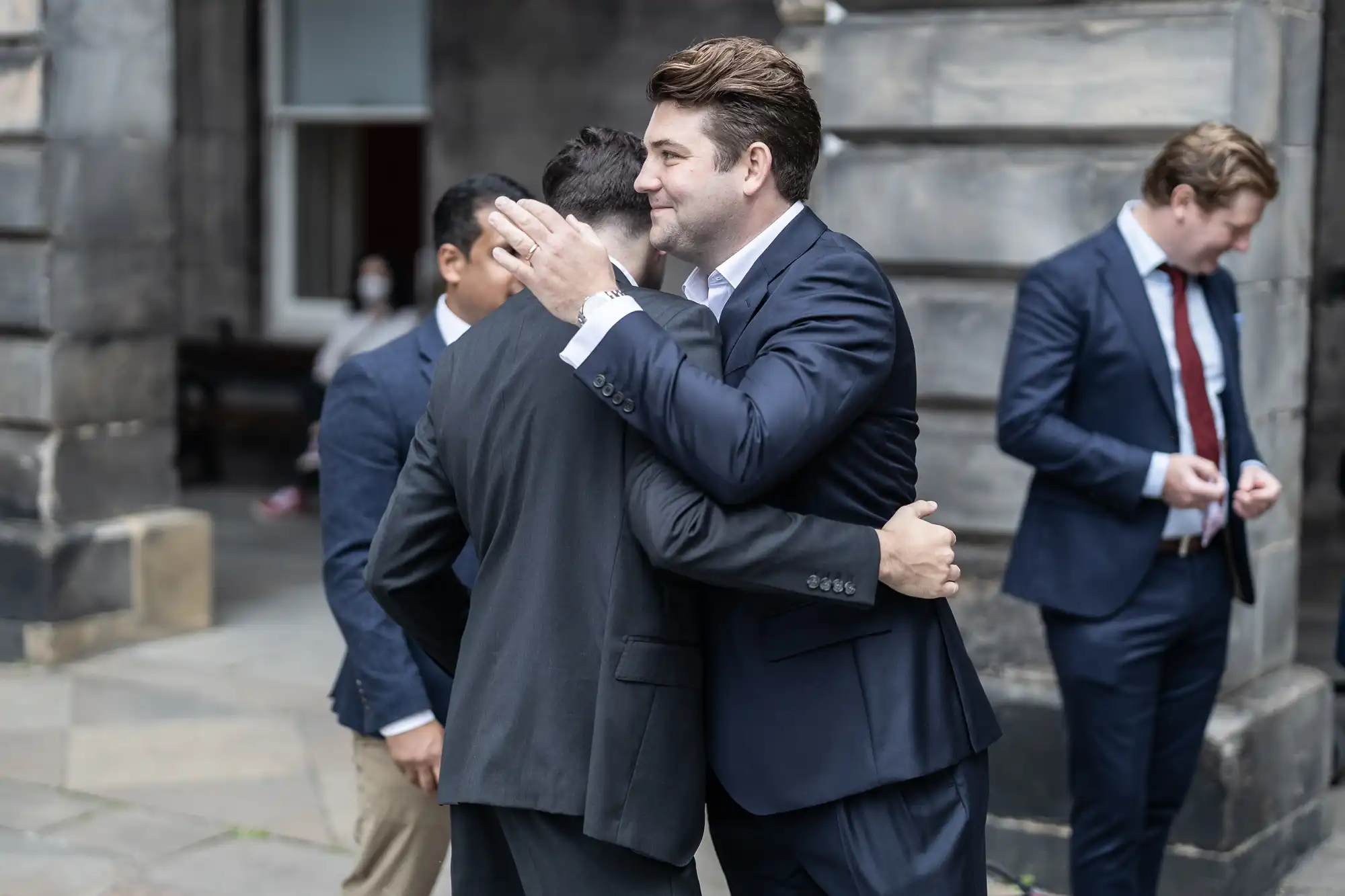 Two men in business suits embracing warmly on a city street, while another man in the background checks his phone.