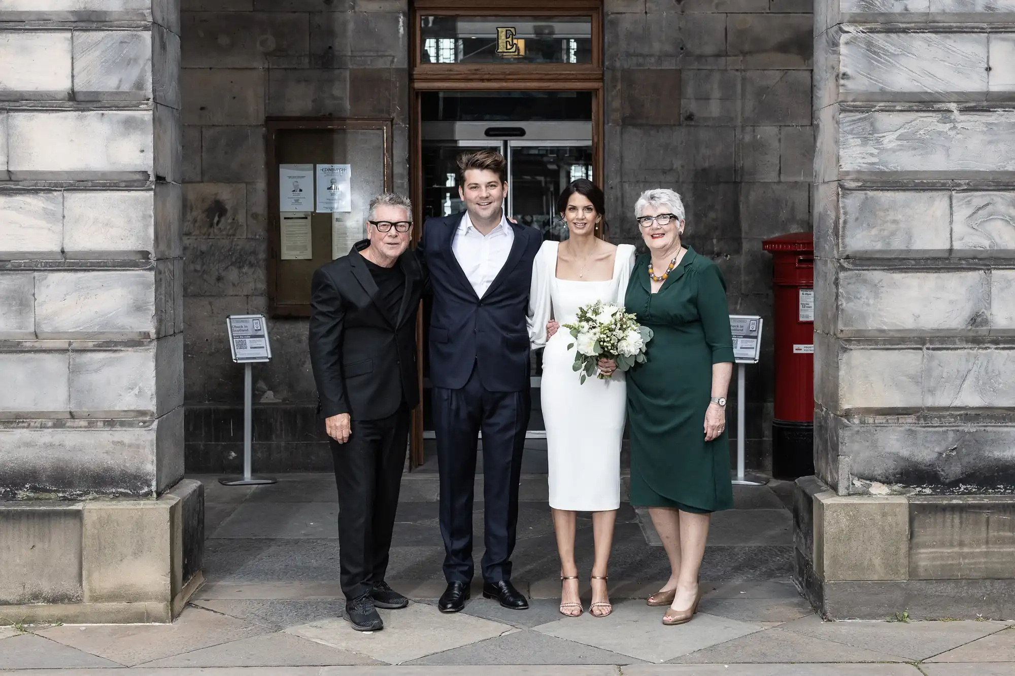 A newlywed couple poses with two older adults outside a stone building, smiling, the bride holding a bouquet.